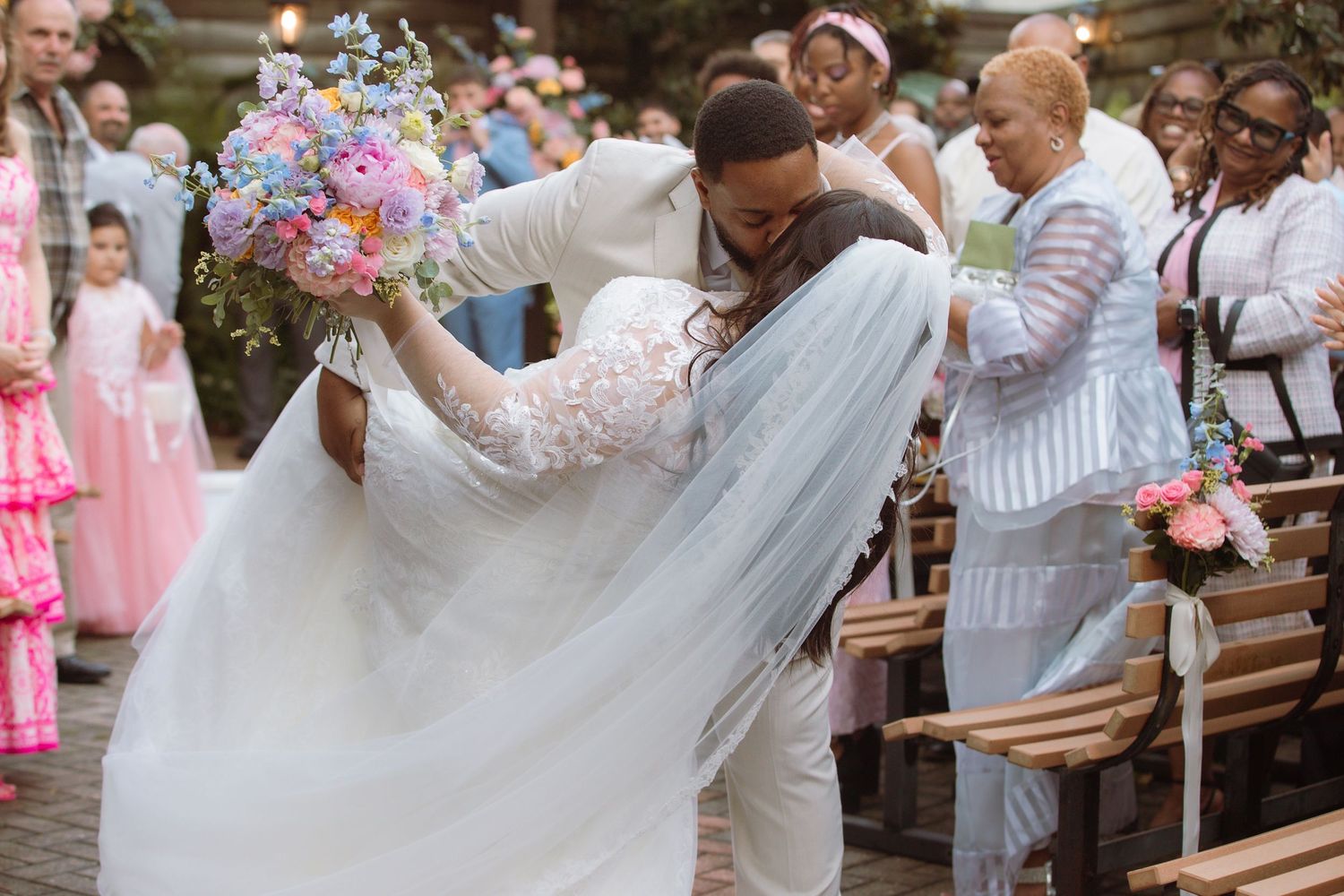 Bride and groom share a romantic dip kiss at outdoor wedding ceremony, colorful bouquet, guests in pink attire.