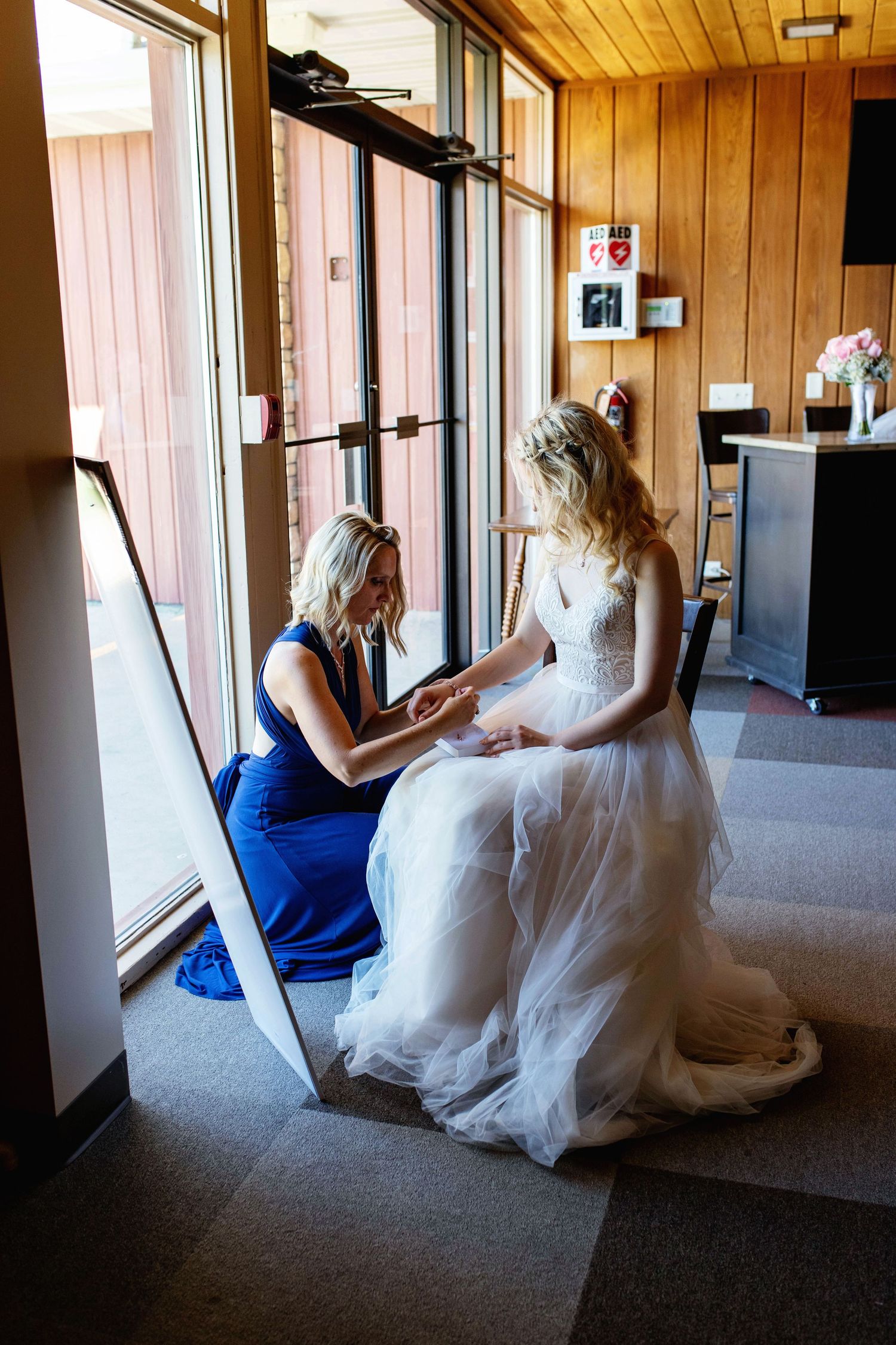 Bridesmaid in blue dress kneels to help bride in white gown with dress details near large windows.