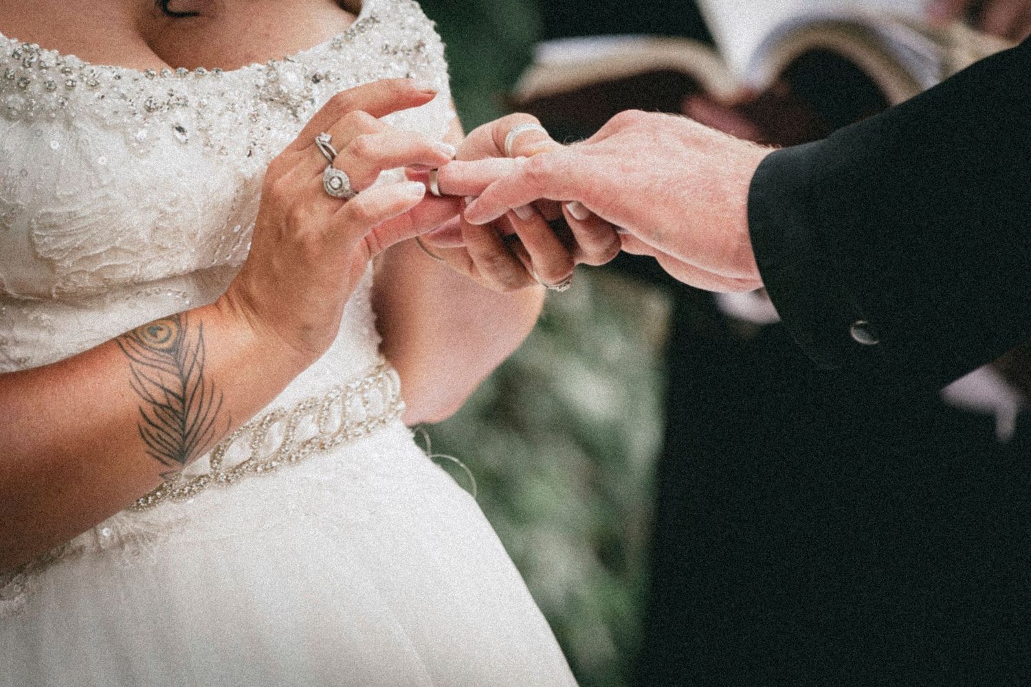 Bride with tattoo placing wedding ring on groom's finger during ceremony, wearing white lace dress.