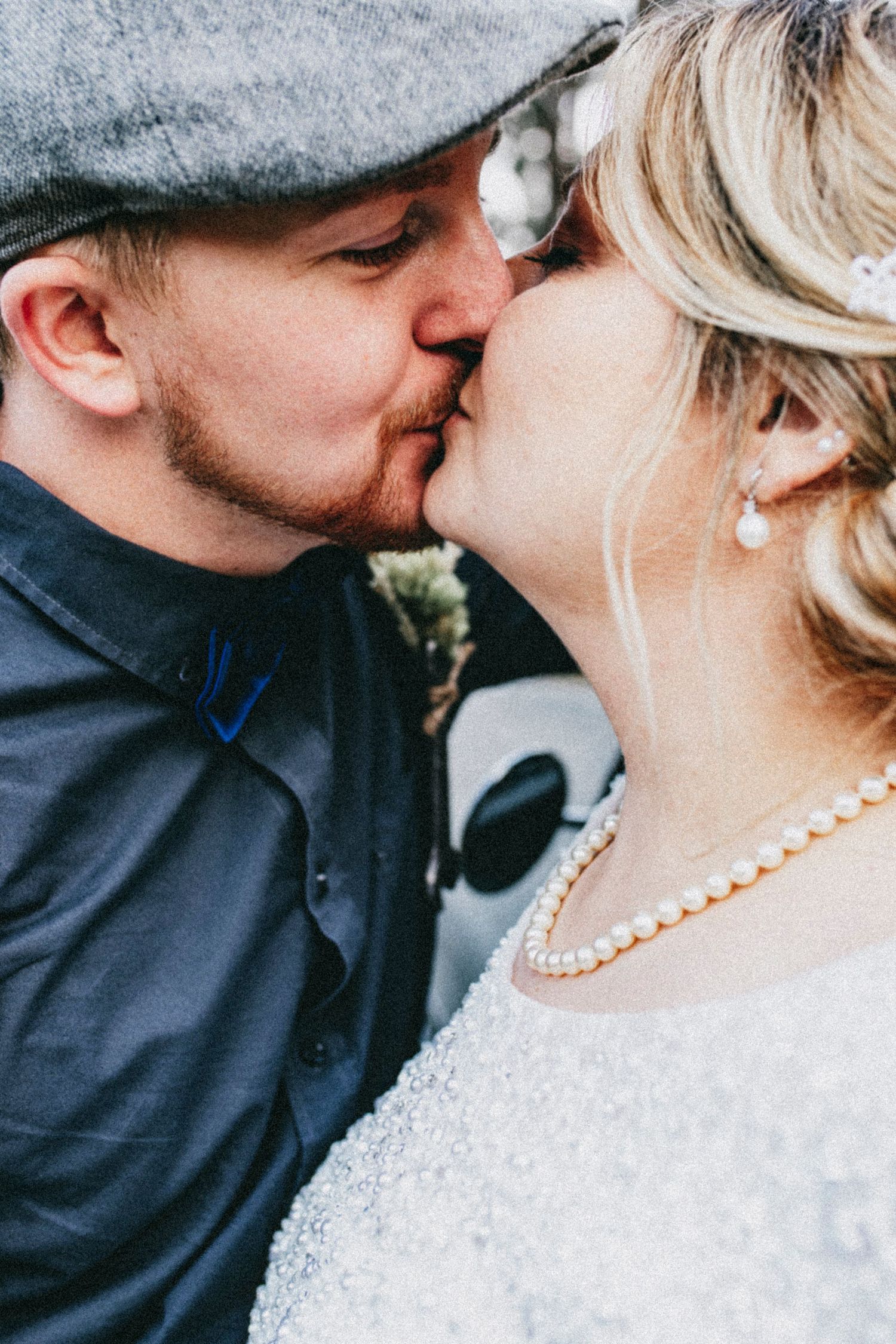 A couple shares a romantic kiss, the groom in a gray flat cap and blue bow tie, the bride wearing a pearl necklace and earrings.