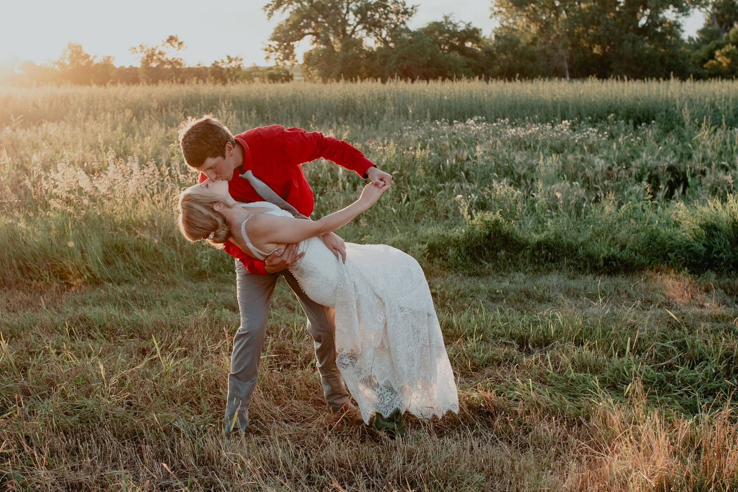 Couple sharing a romantic dip kiss in a golden sunlit field, groom in red jacket, bride in white wedding dress.