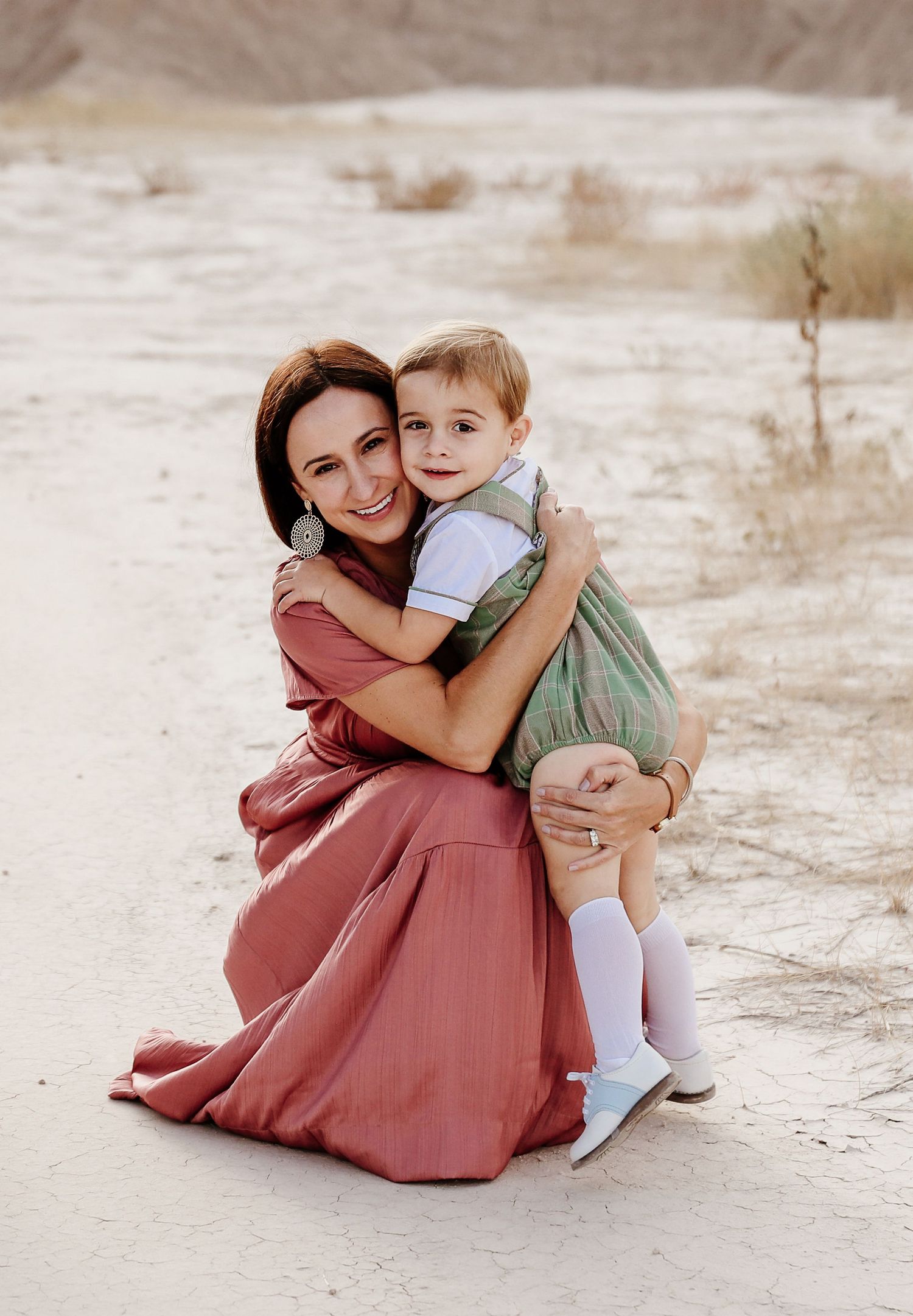 Mother in pink maxi dress hugging young son in green romper, smiling outdoors in desert setting.