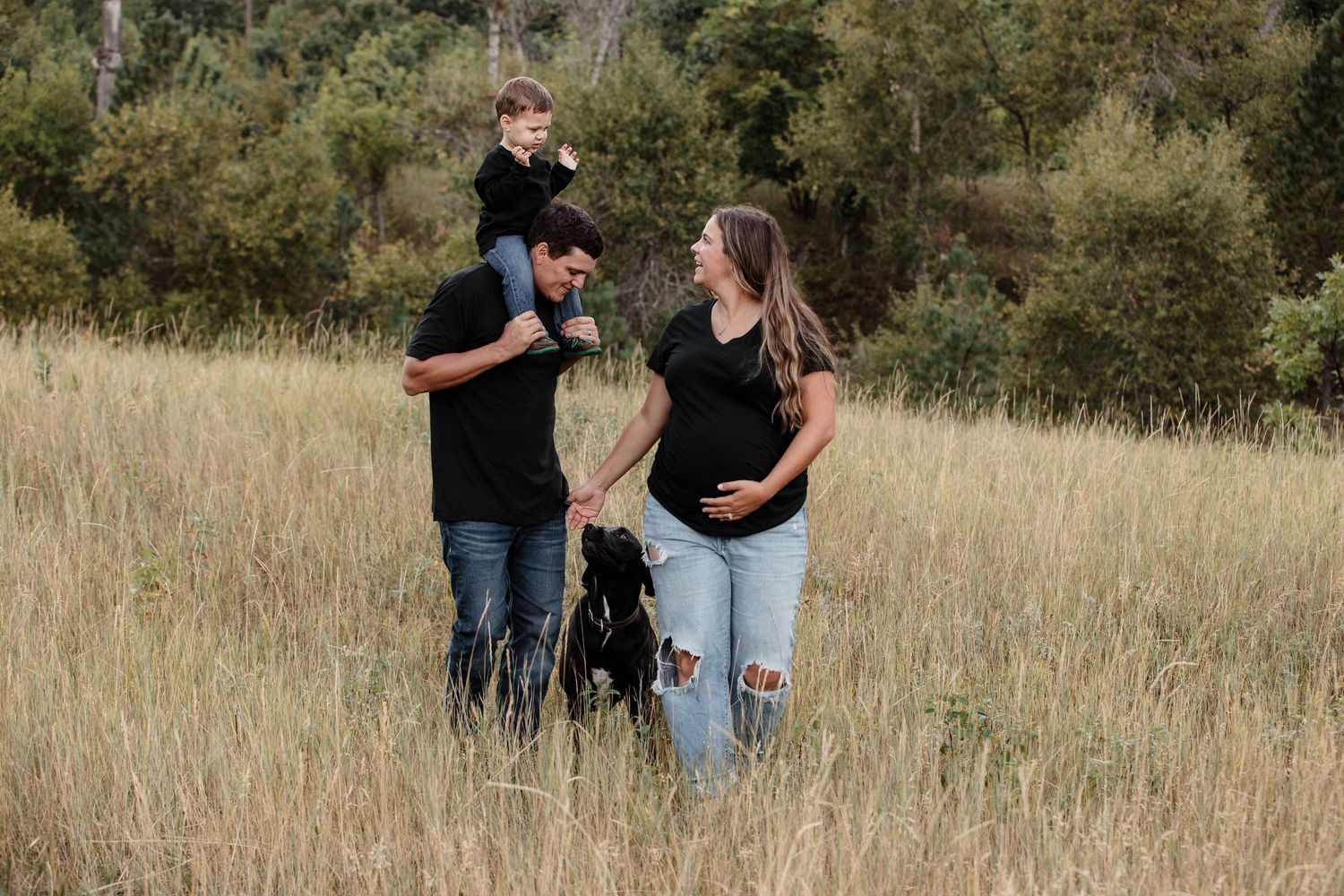 Couple in black with toddler on shoulders and dog poses in tall golden grass field.