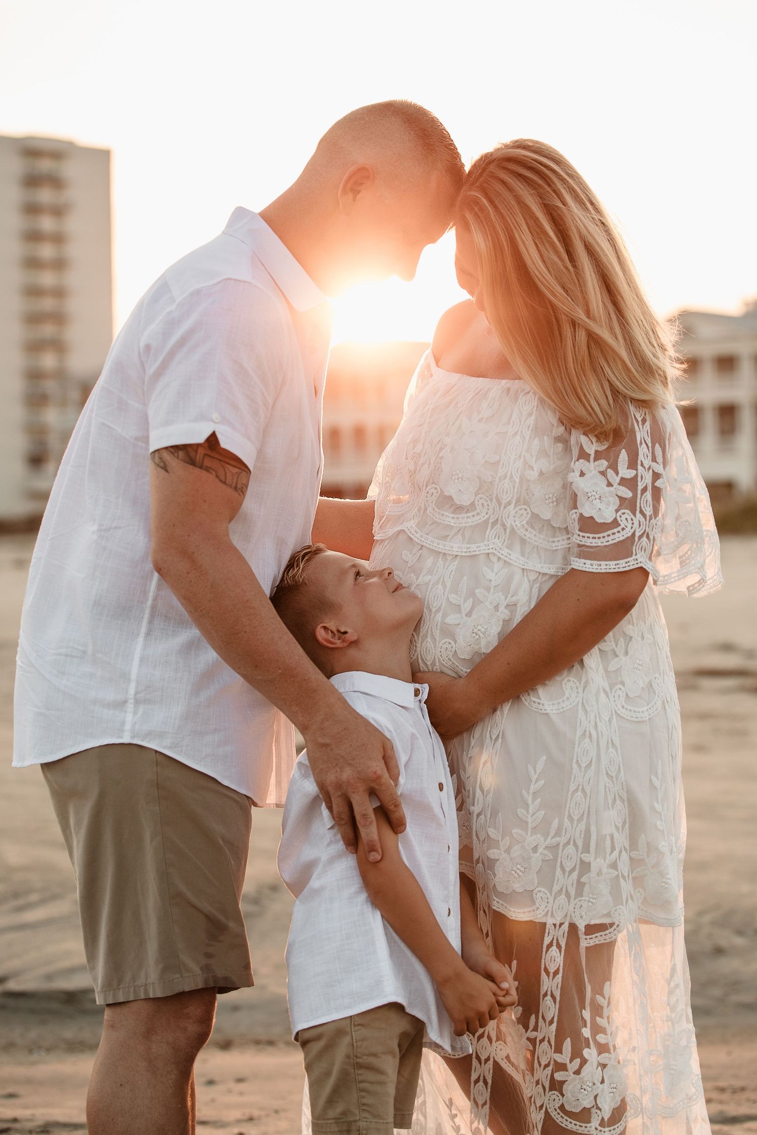 Family of three posing at sunset on beach, parents touching foreheads while son looks up between them.