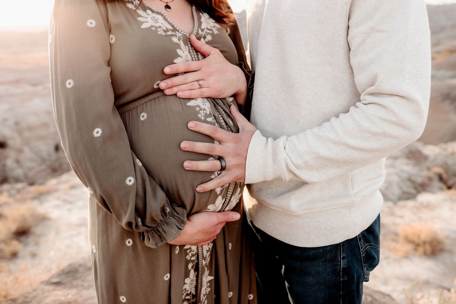 Couple cradles pregnant belly together outdoors in desert landscape at golden hour.