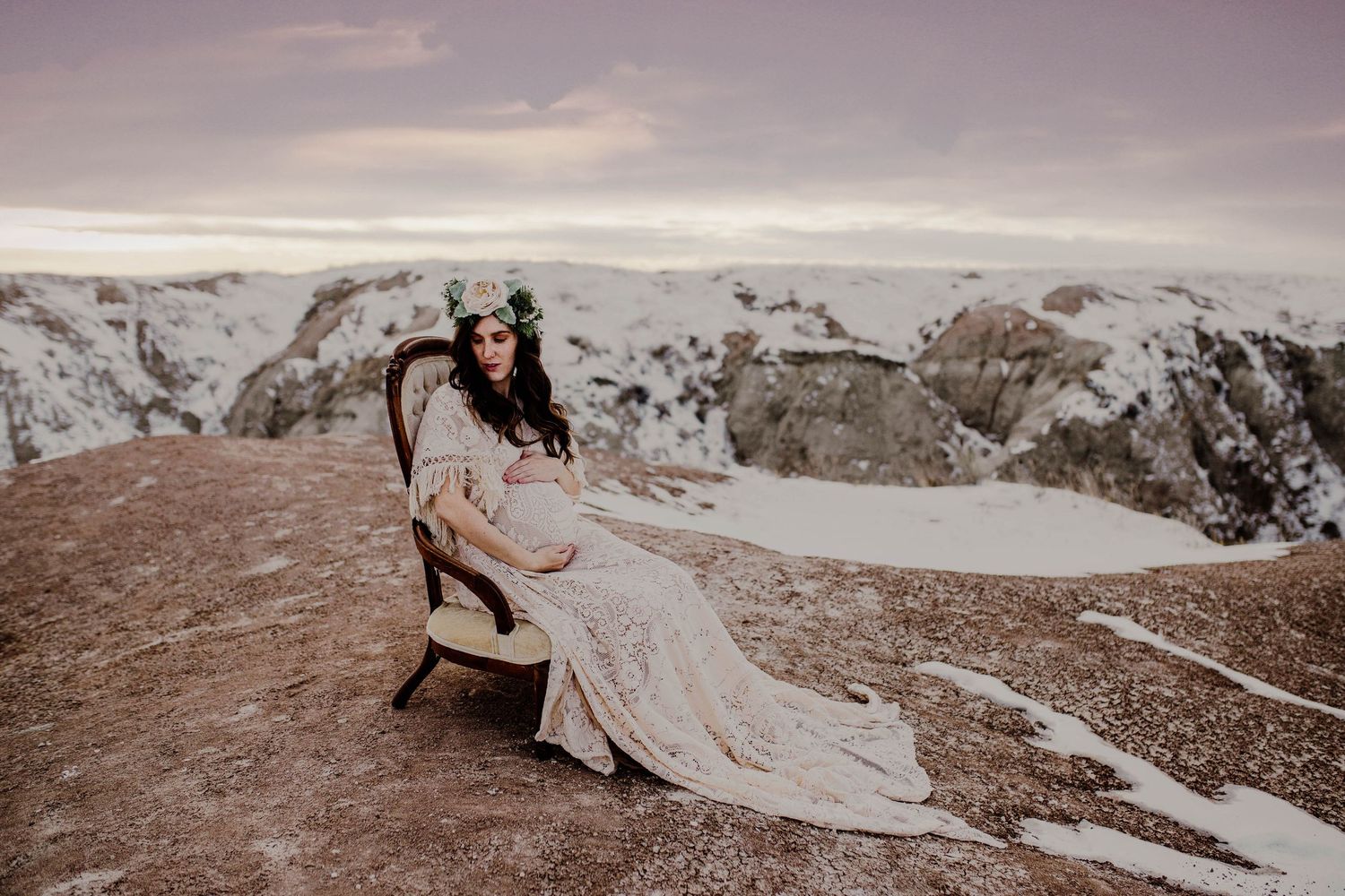Pregnant woman in lace gown and floral crown sits in vintage chair on snowy mountain overlook.