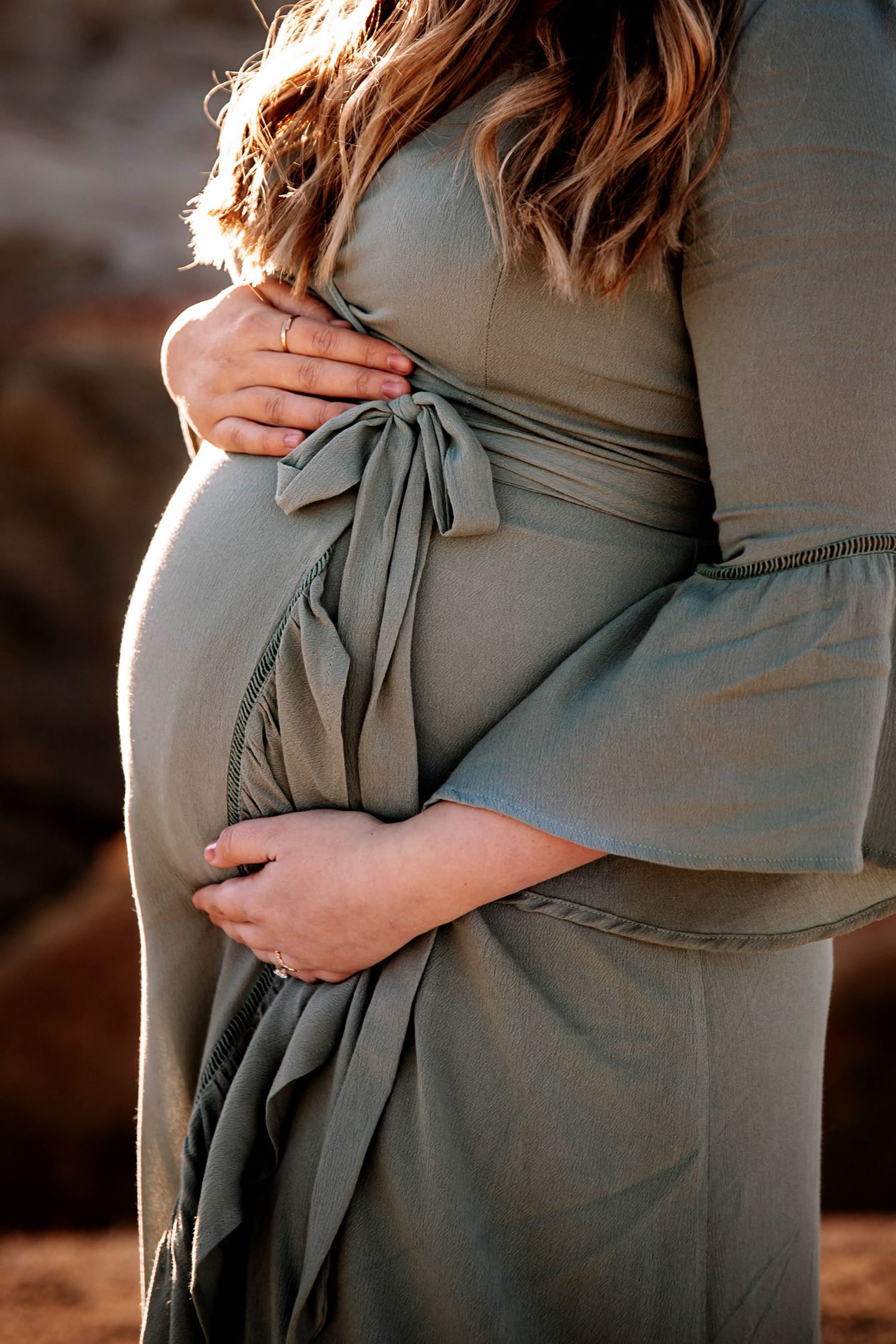 Close-up of pregnant woman in sage green wrap dress cradling baby bump outdoors.