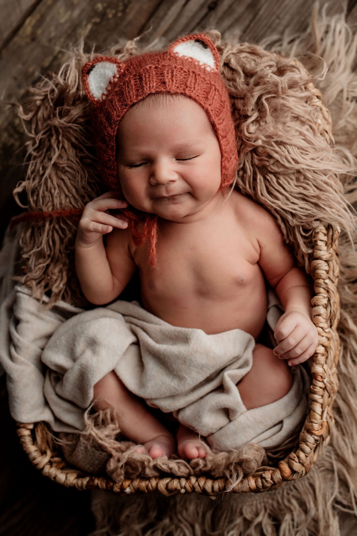 Smiling newborn baby wearing knit fox hat resting in wicker basket with blanket.