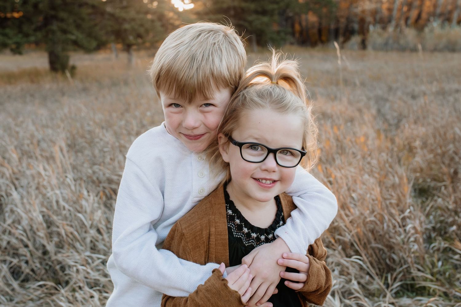 Young boy hugging his little sister wearing glasses in a warm autumn field during a family photo session.
