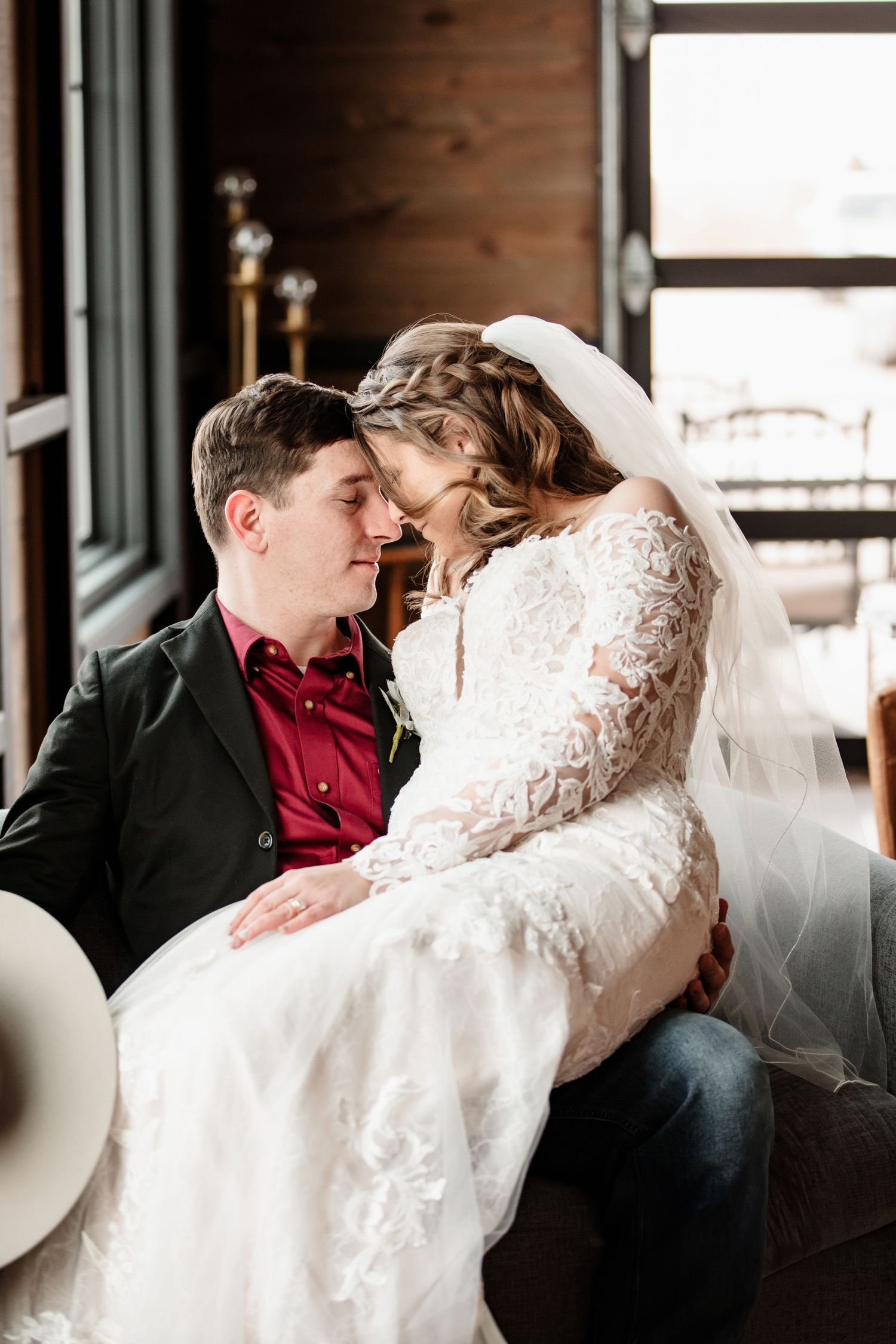 Bride in lace gown and veil sits on groom's lap in intimate forehead-to-forehead pose during wedding portrait.
