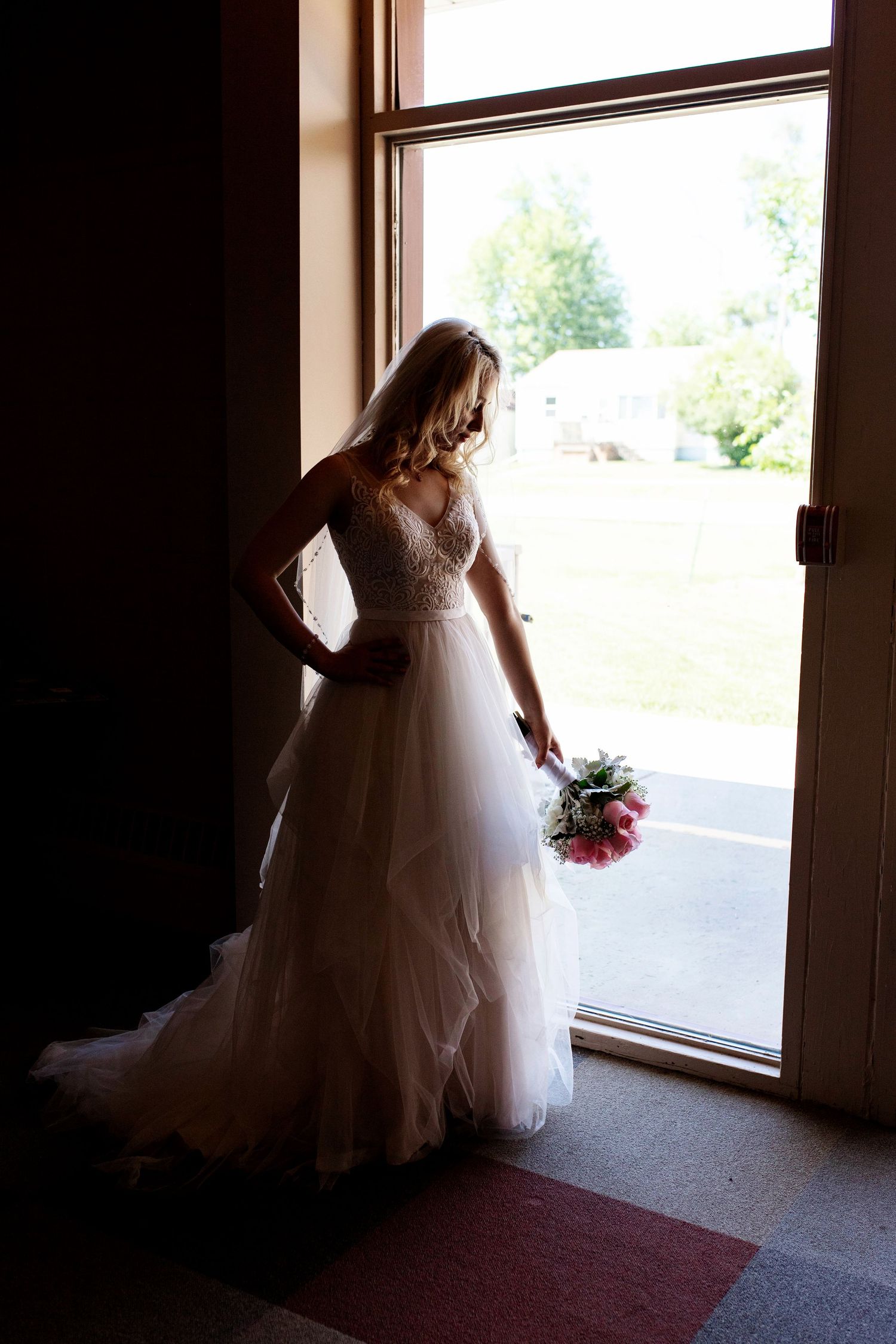 Bride in white tulle gown holding pink bouquet stands in doorway, backlit by natural light.