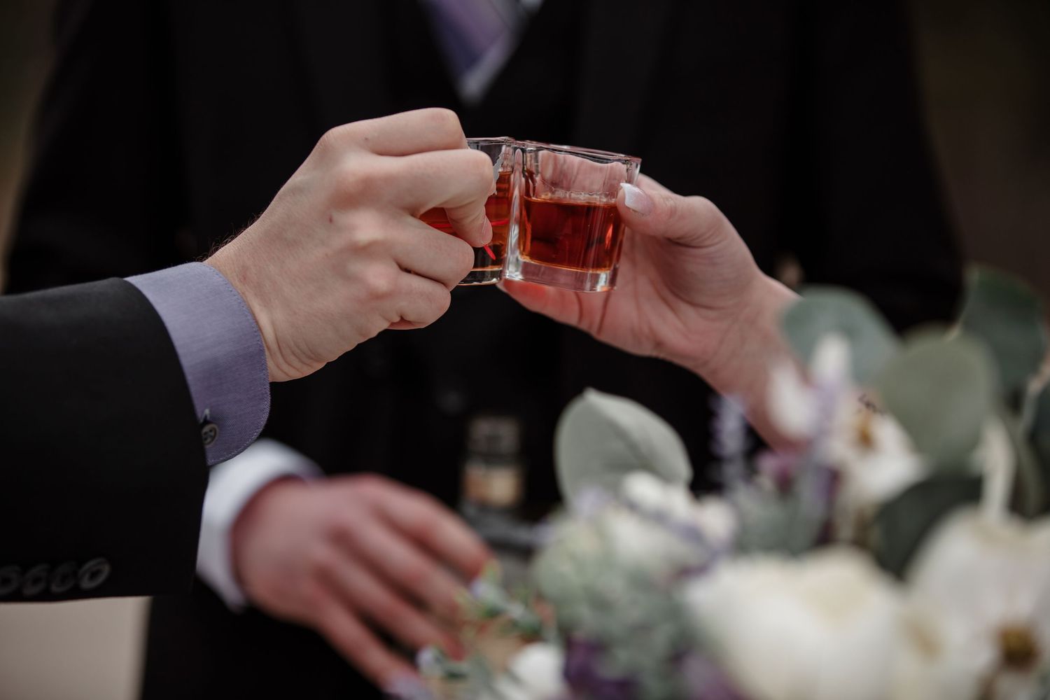 Two men in suits clinking small glasses of amber liquor at a wedding celebration with floral arrangements.