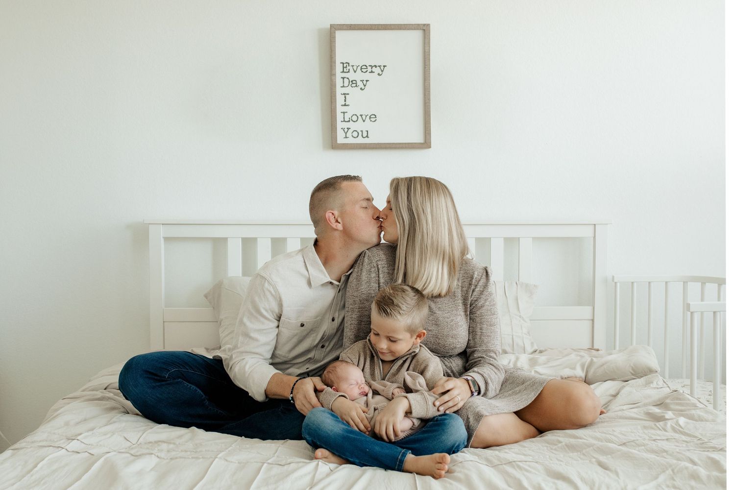 A family shares a tender moment together while sitting on a white bed beneath a simple framed artwork.