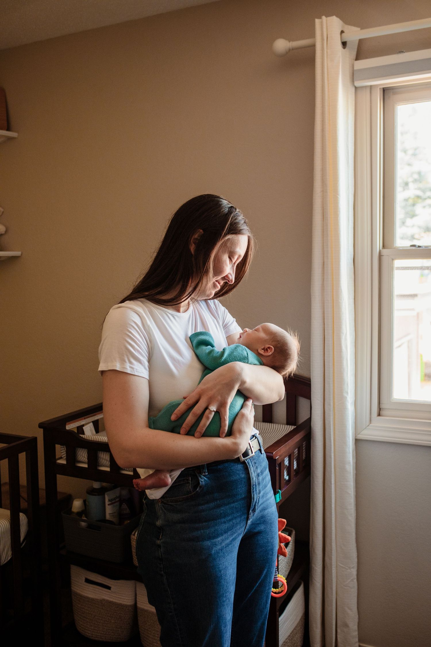 A caring parent holds and looks down lovingly at a swaddled newborn baby near a sunlit window in a nursery.