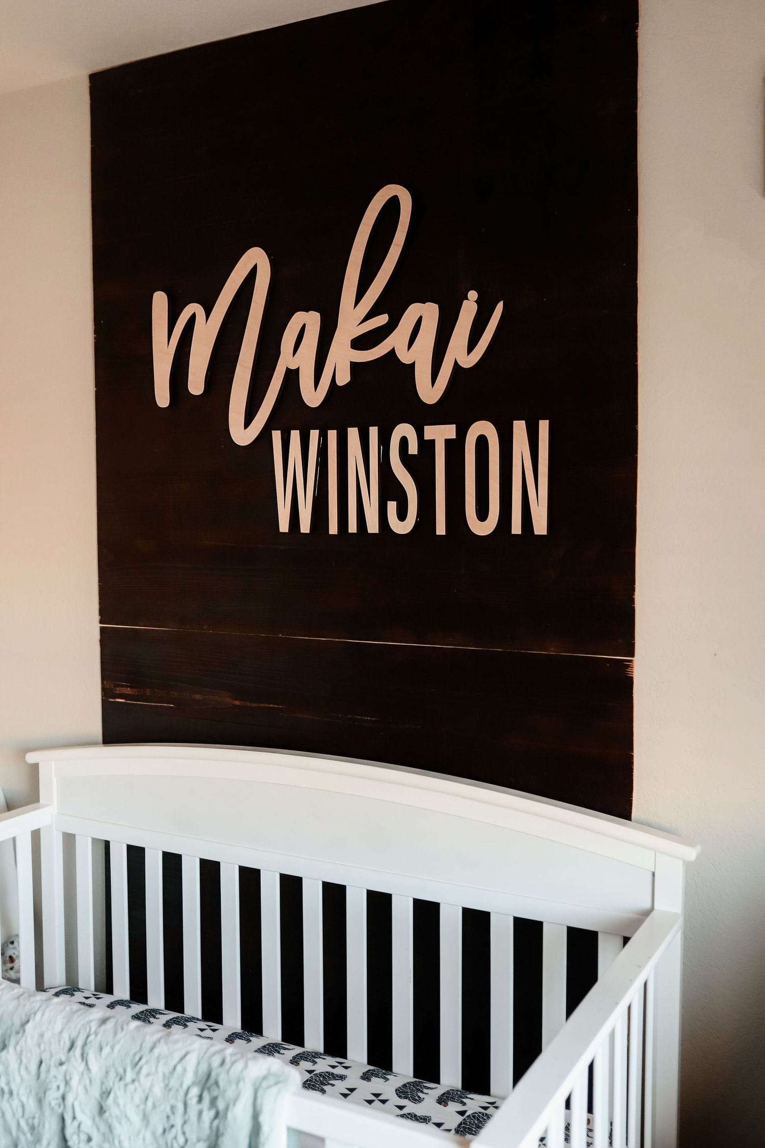 Dark wood nursery name sign reading Makai Winston in pink lettering mounted above white baby crib.