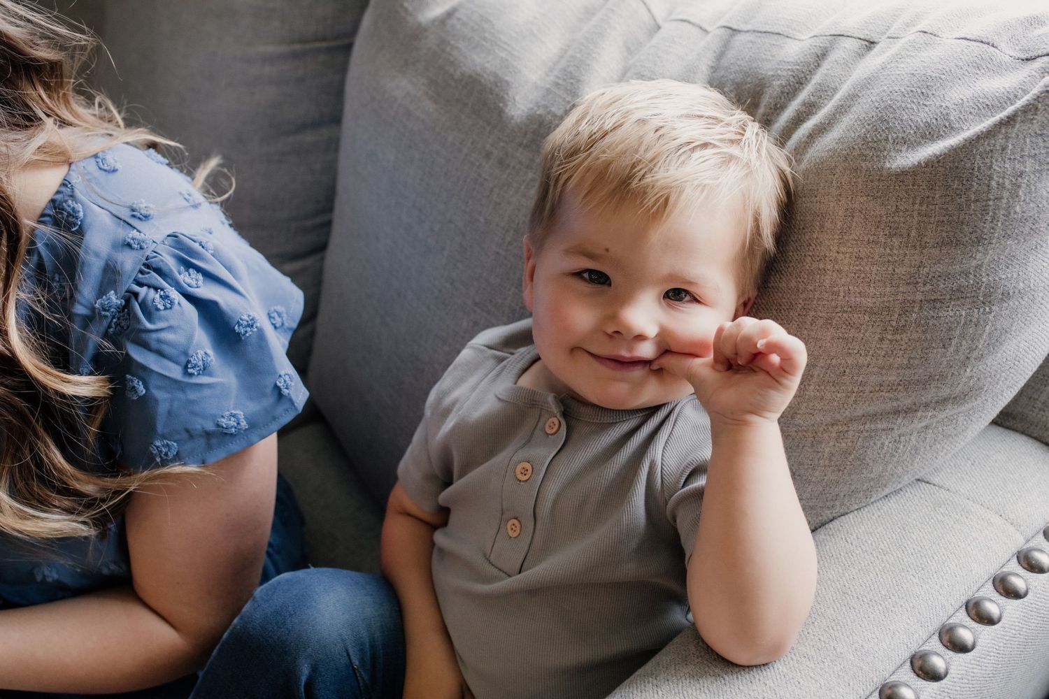 Adorable smiling toddler boy in gray shirt sitting on a gray couch looking at the camera.
