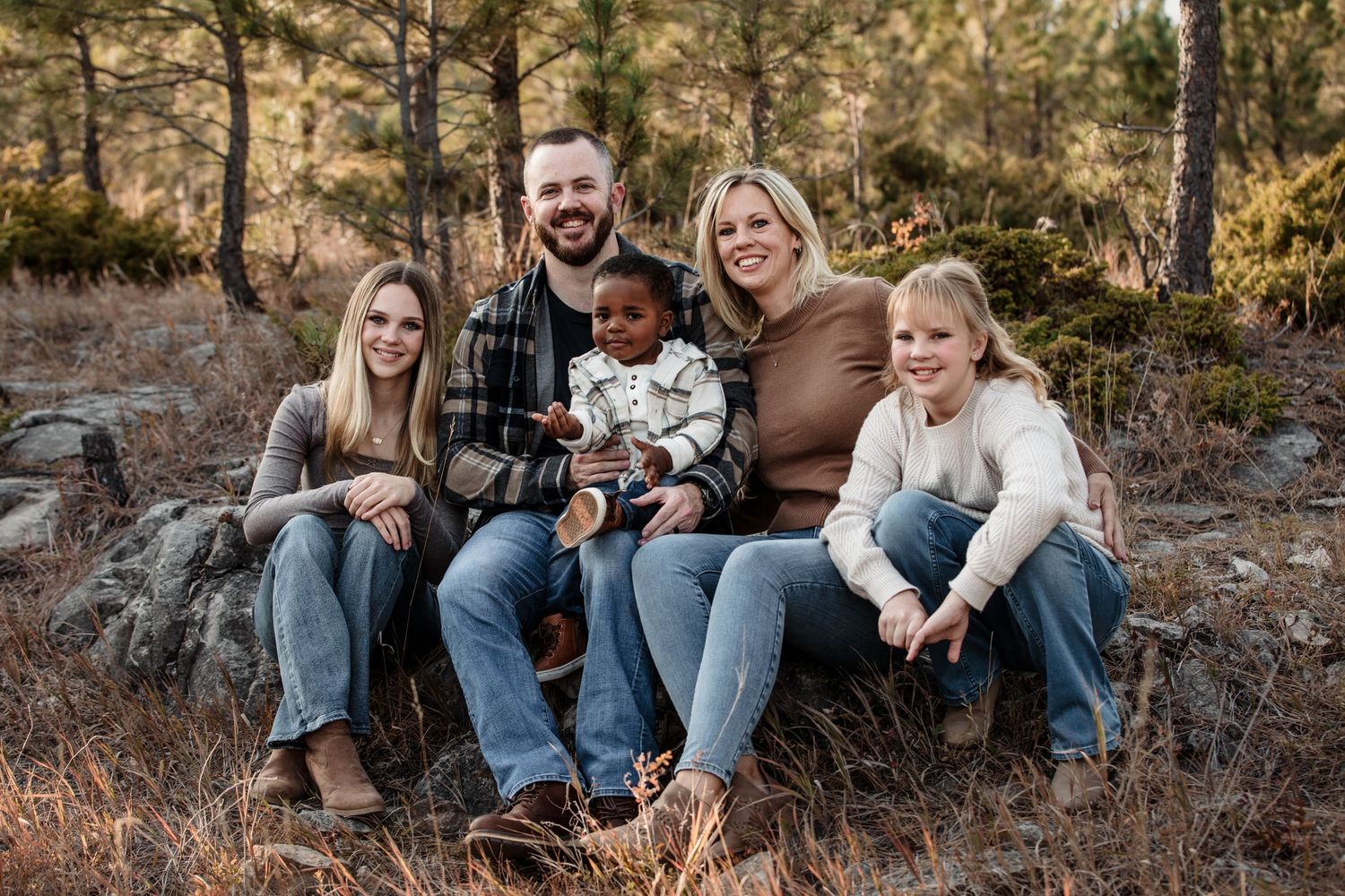 Happy family of five posing outdoors in a forest setting, smiling together on a rustic log in casual attire.