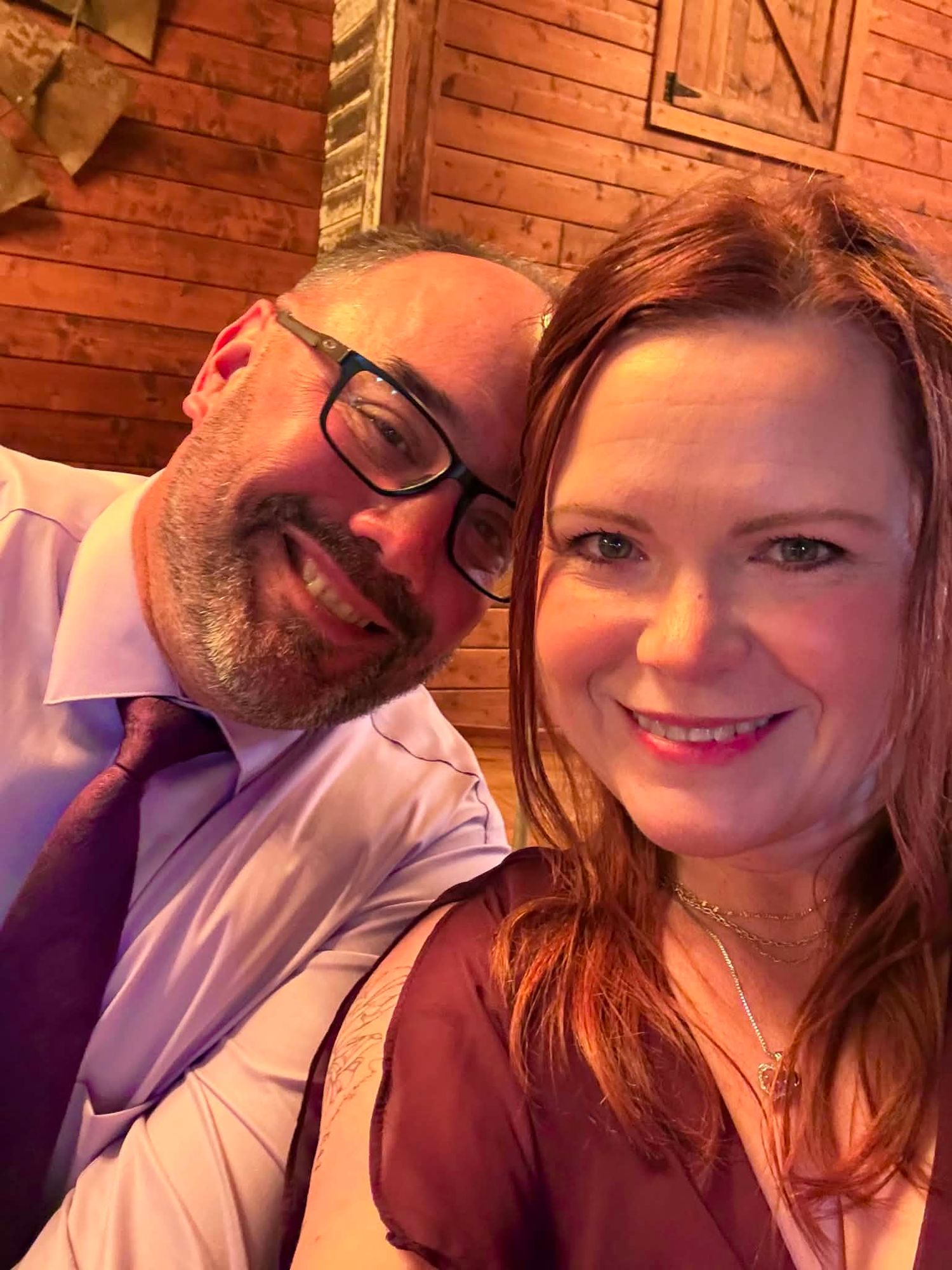 A smiling couple poses for a selfie, man with glasses and beard in purple shirt, woman with red hair in warm-lit rustic setting.