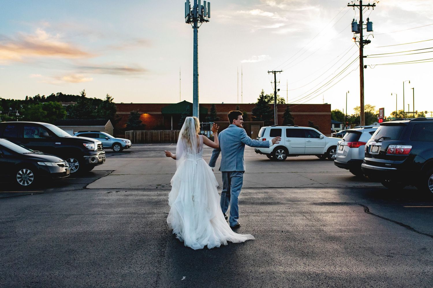 Bride and groom walking hand in hand through a parking lot at sunset, bride wearing a flowing white gown.