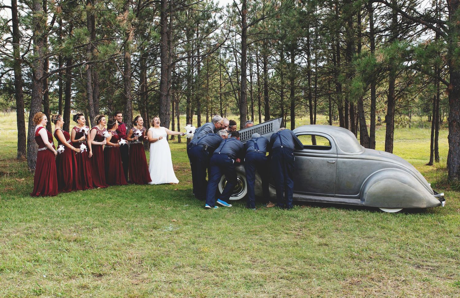 Wedding party poses beside a vintage gray coupe in a pine forest, bridesmaids in burgundy dresses and groomsmen inspecting the classic car.