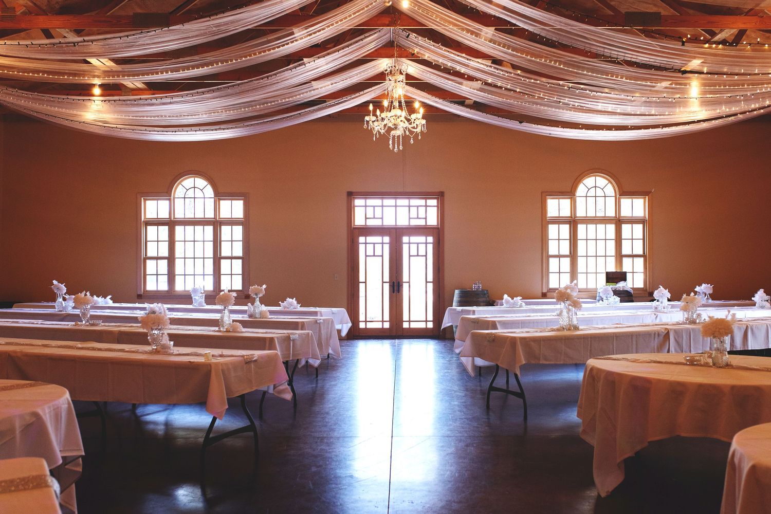 Elegant banquet hall with white draped ceiling, chandelier, arched windows, and white linen-covered tables set for a formal event.