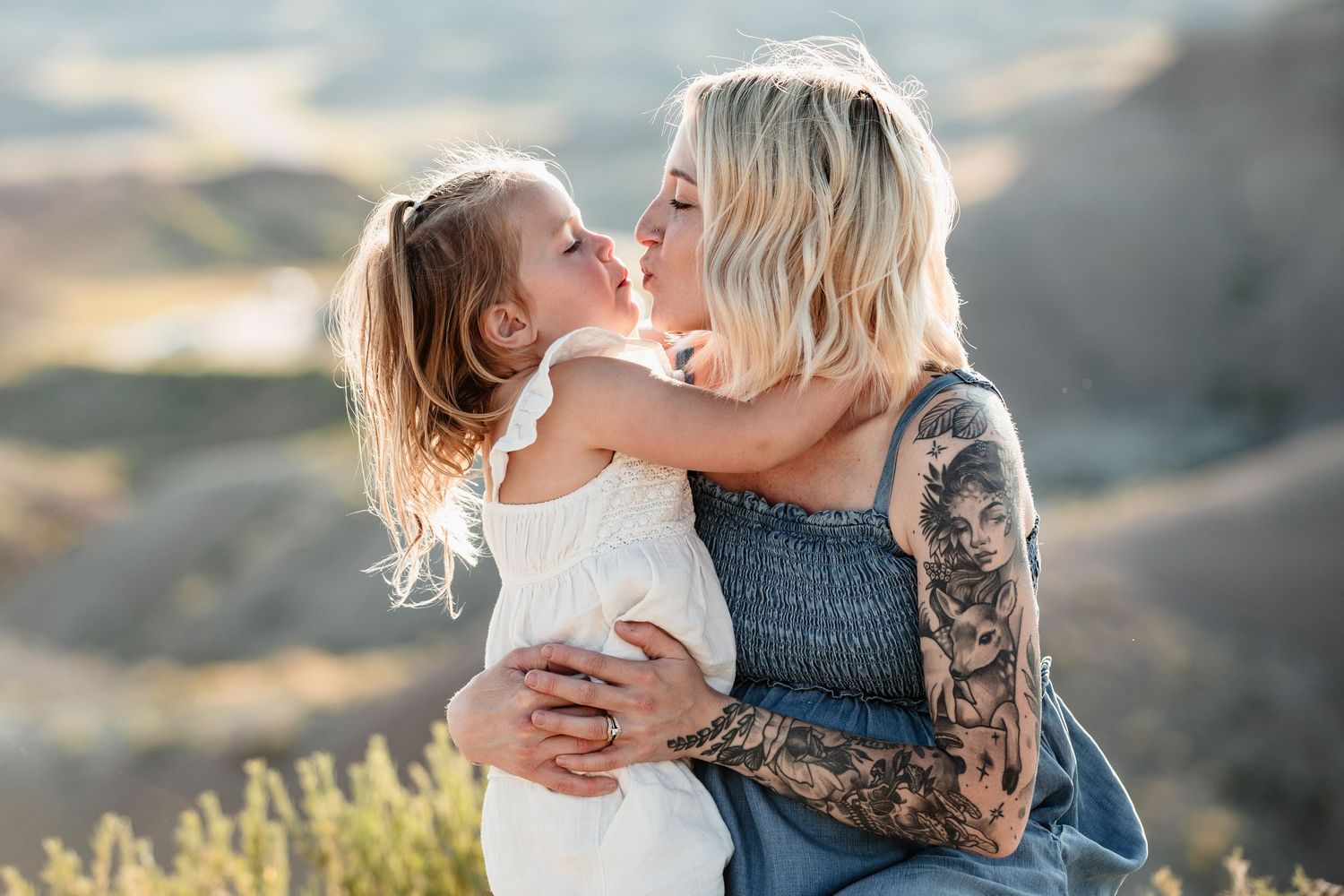 A tattooed blonde mother lovingly kisses her young daughter outdoors in a scenic desert landscape.