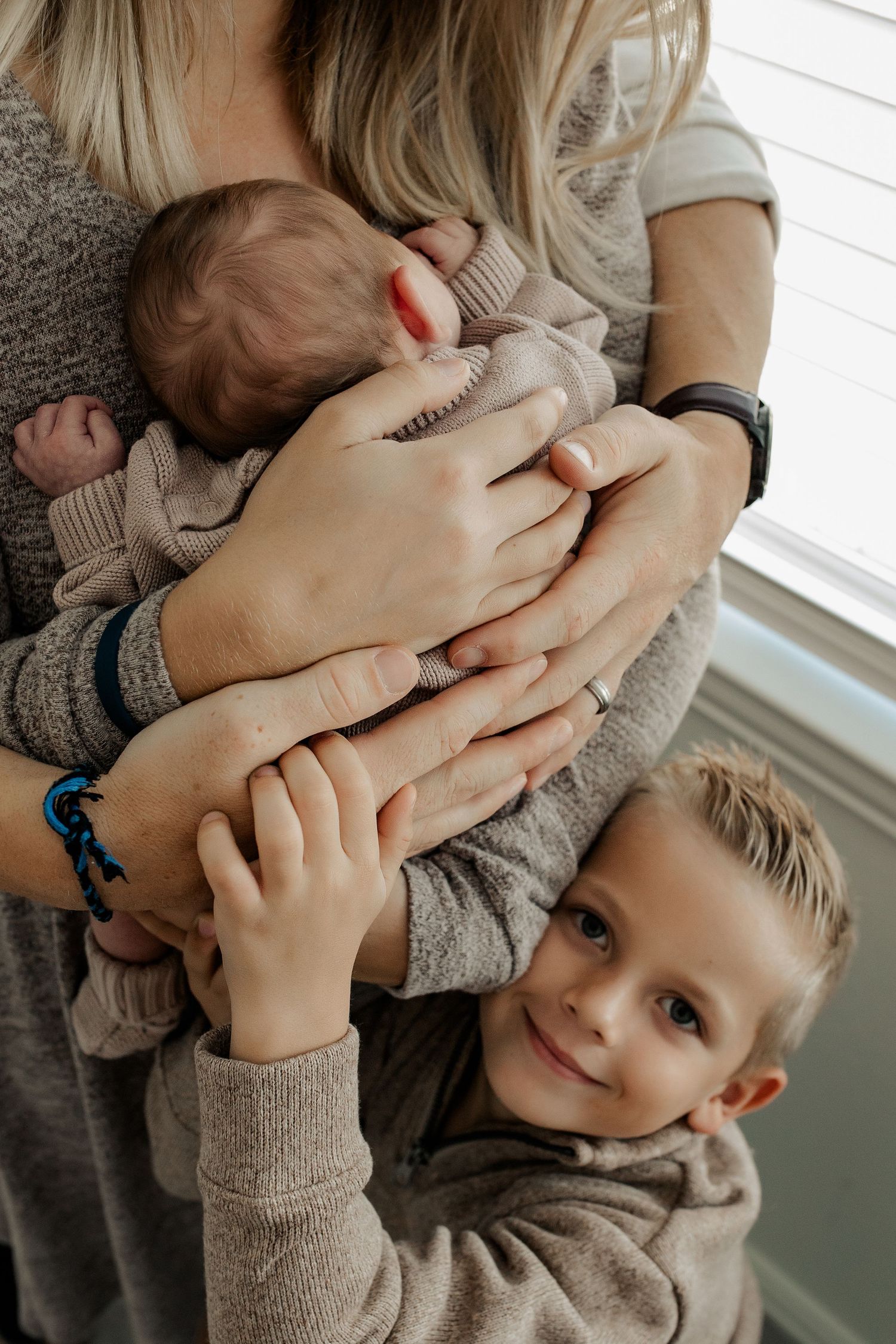 Young boy smiling up at camera while hugging mother holding newborn baby sibling near bright window.