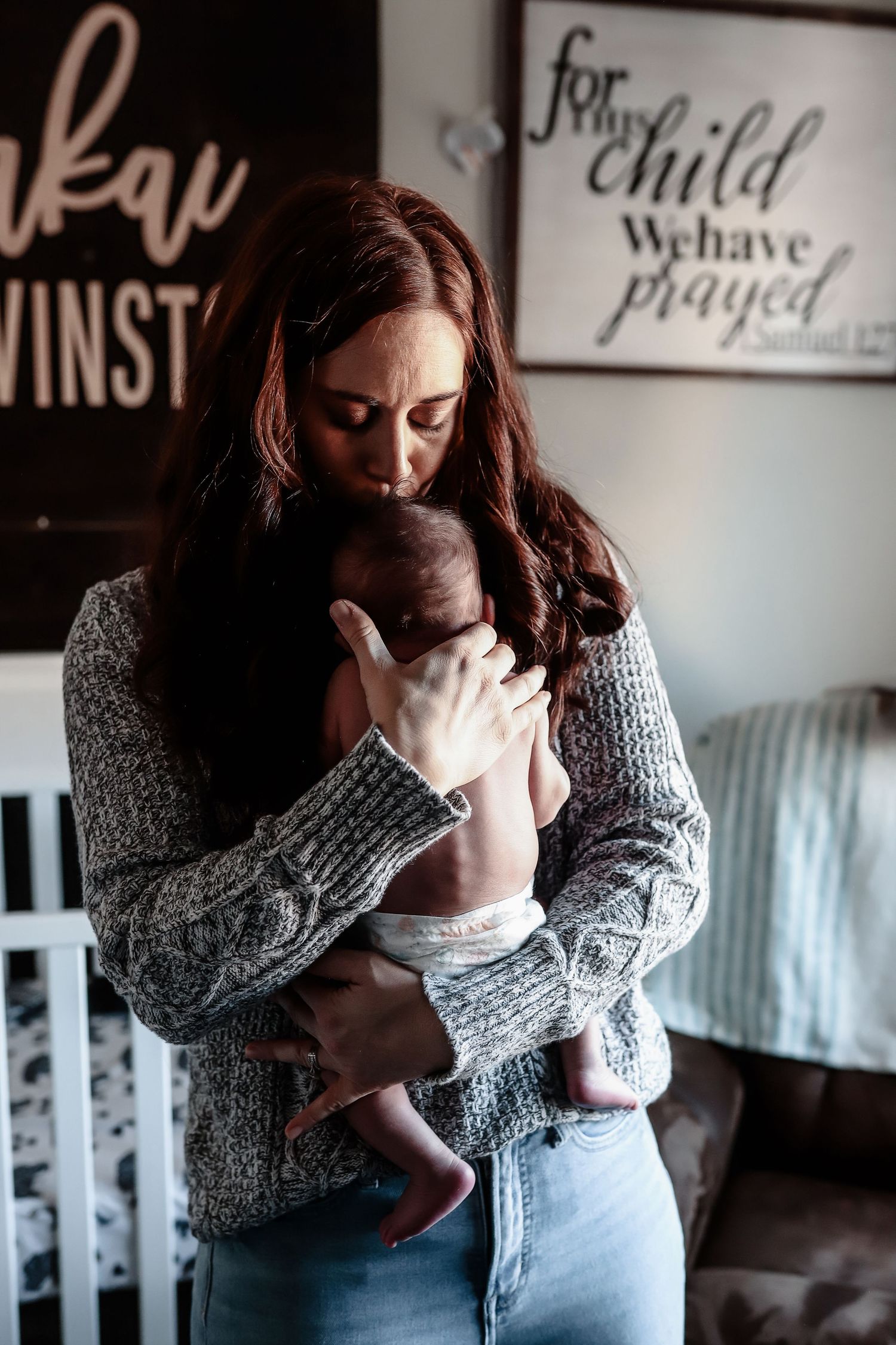 Red-haired mother gently kisses her newborn baby in a nursery with inspirational wall signs.