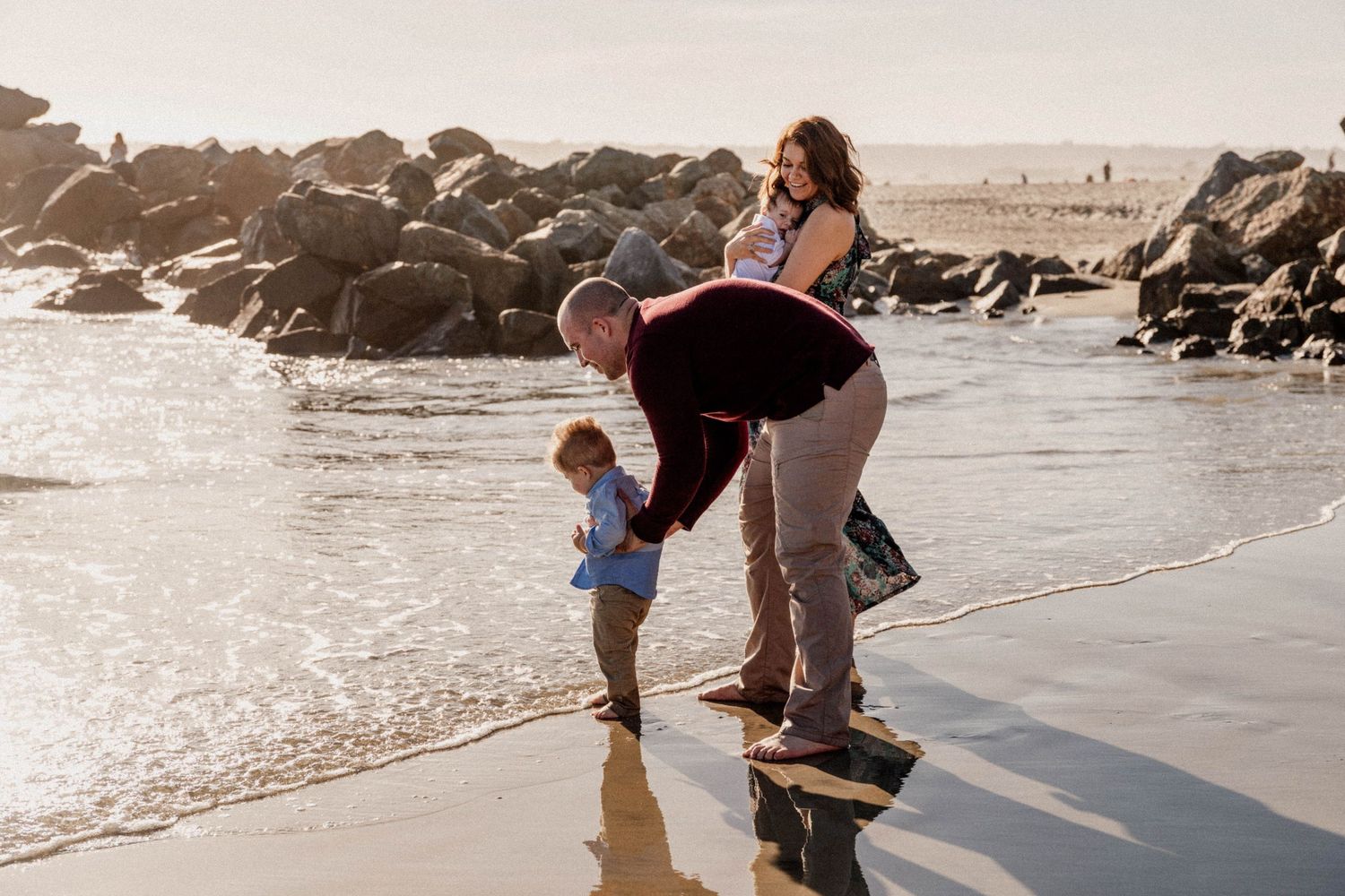 Father helping toddler touch ocean waves at beach while mother watches, rocks in background.