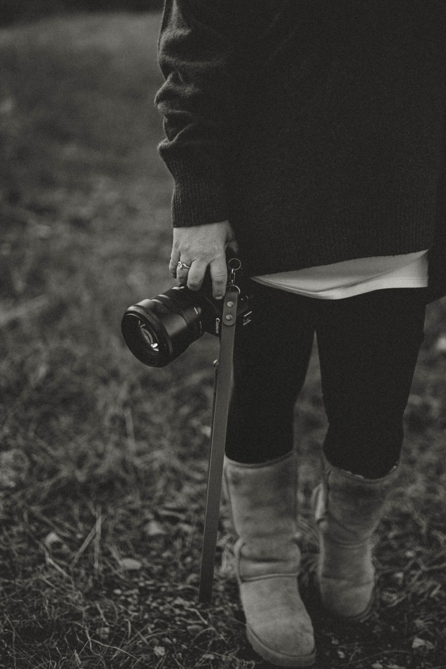 Black and white close-up of a photographer holding a camera with UGG boots in a natural outdoor setting.