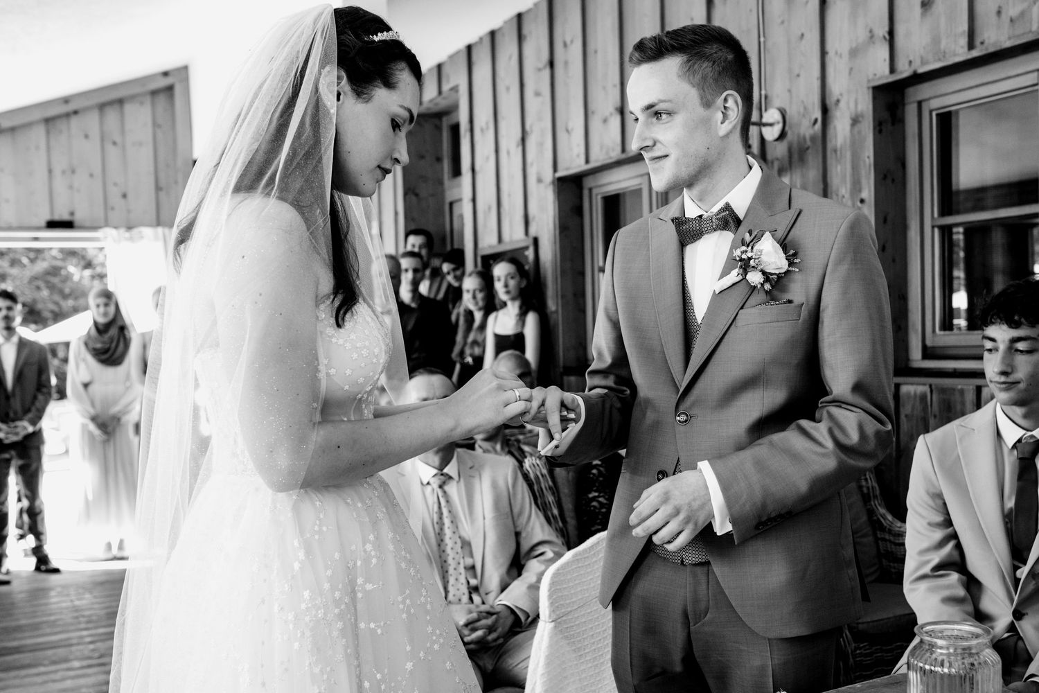 Bride placing wedding ring on groom's finger during outdoor ceremony, both smiling in black and white photo.