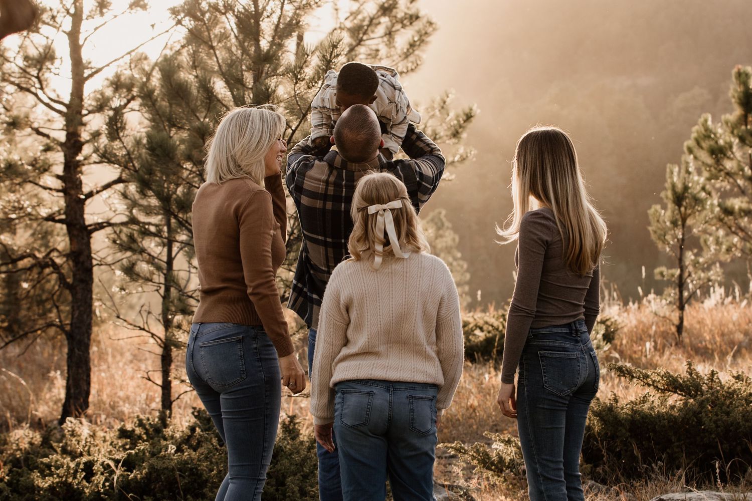 Family of four standing together outdoors in a sunlit forest, viewed from behind, wearing casual autumn clothing.