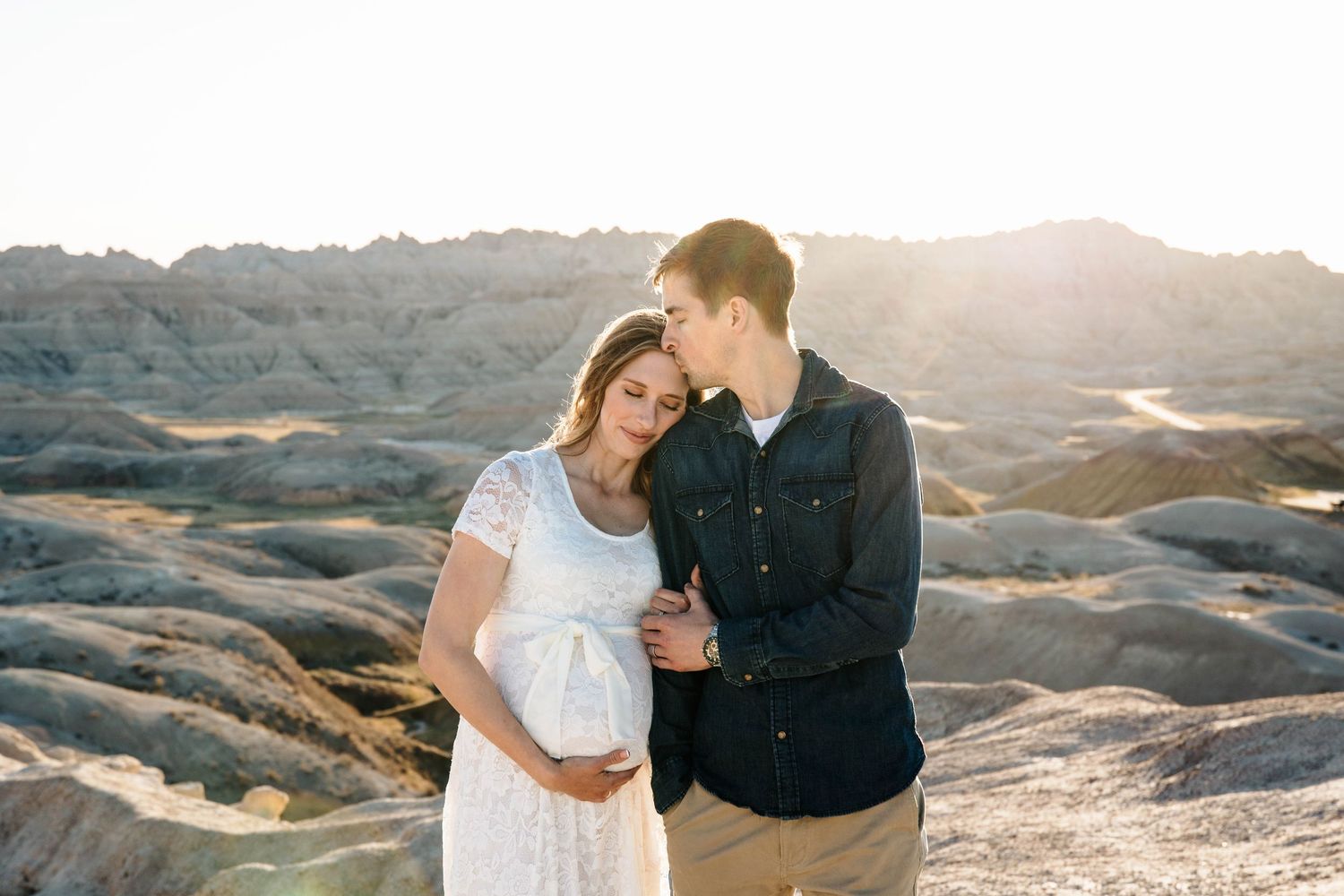 Couple embracing at sunset in the Badlands, pregnant woman in white lace dress with partner.
