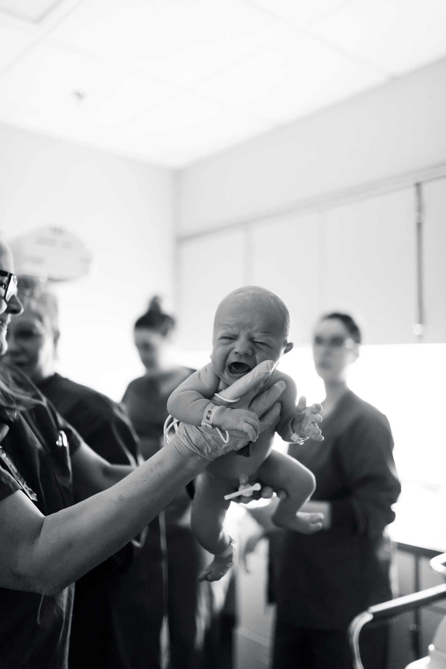 Medical professional lifts crying newborn baby in delivery room with staff in background, black and white.