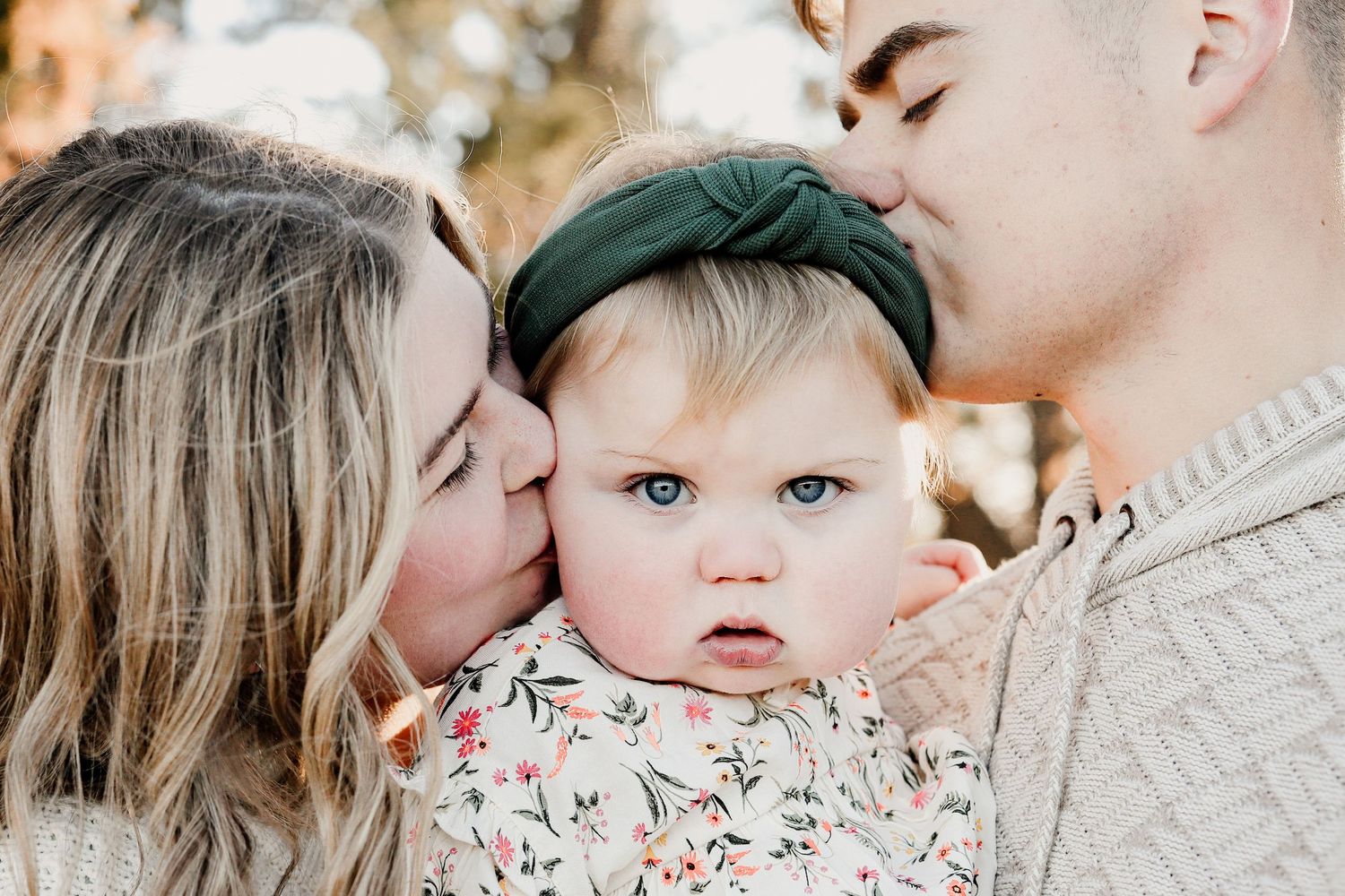 Two parents lovingly kissing their blue-eyed baby girl on the cheeks in a tender family portrait.