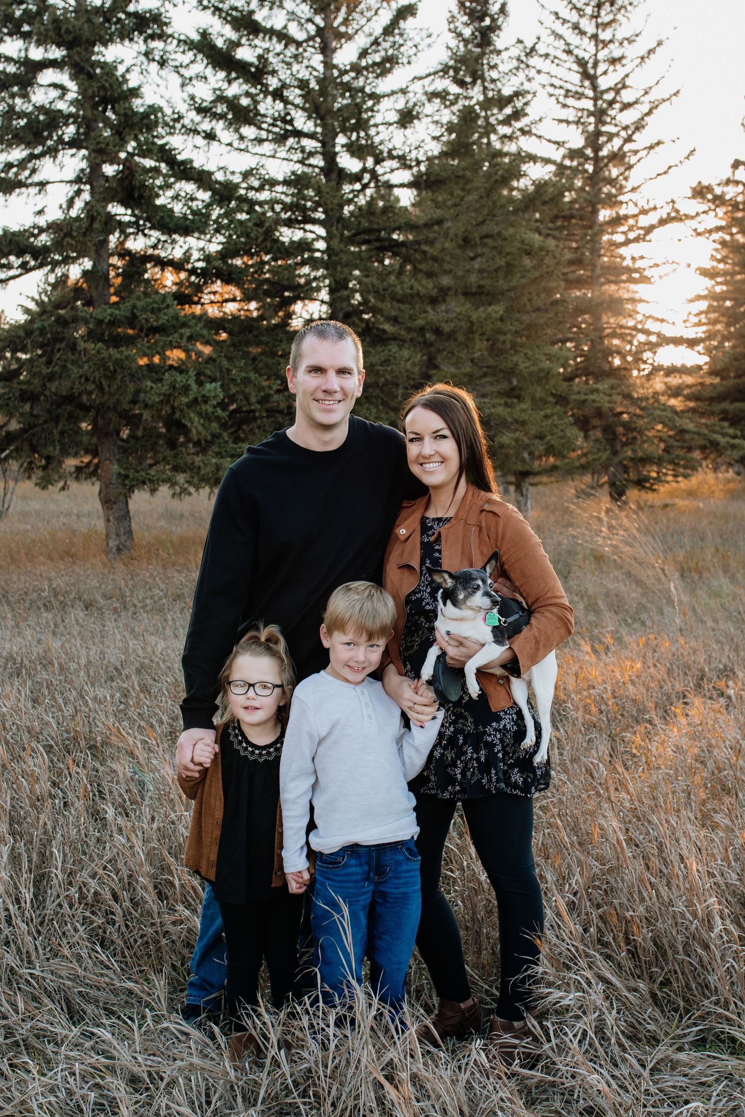 Happy family of four with a small dog posing in a golden field surrounded by pine trees at sunset.