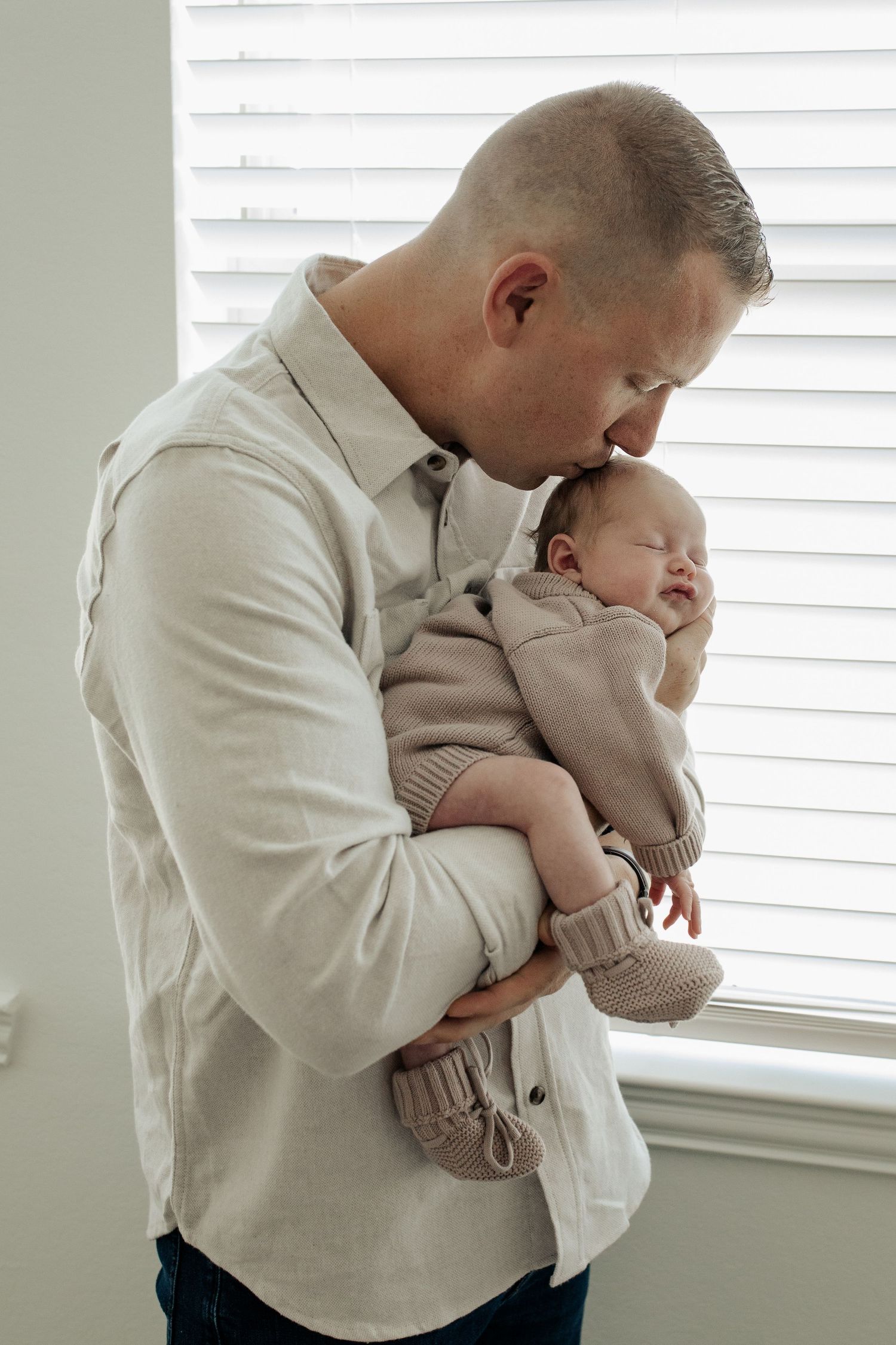 Father in beige shirt gently kissing sleeping newborn baby held in arms near bright window with blinds.
