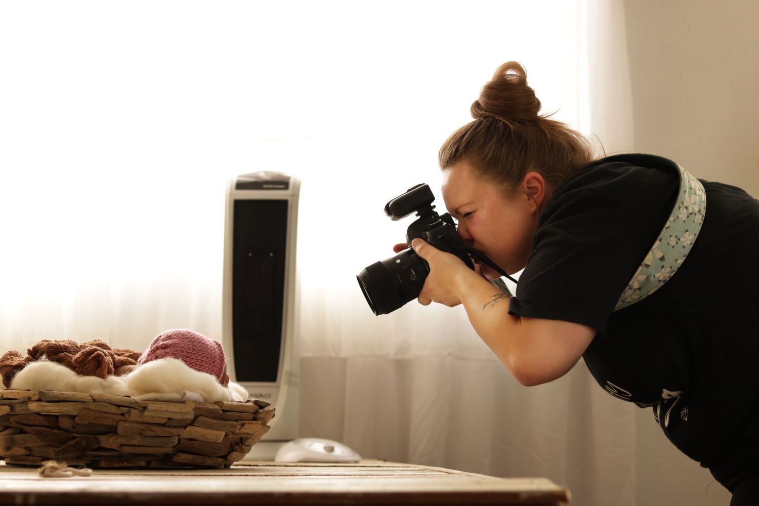 Photographer in black shirt captures close-up shots of fruit arranged in woven basket near window.