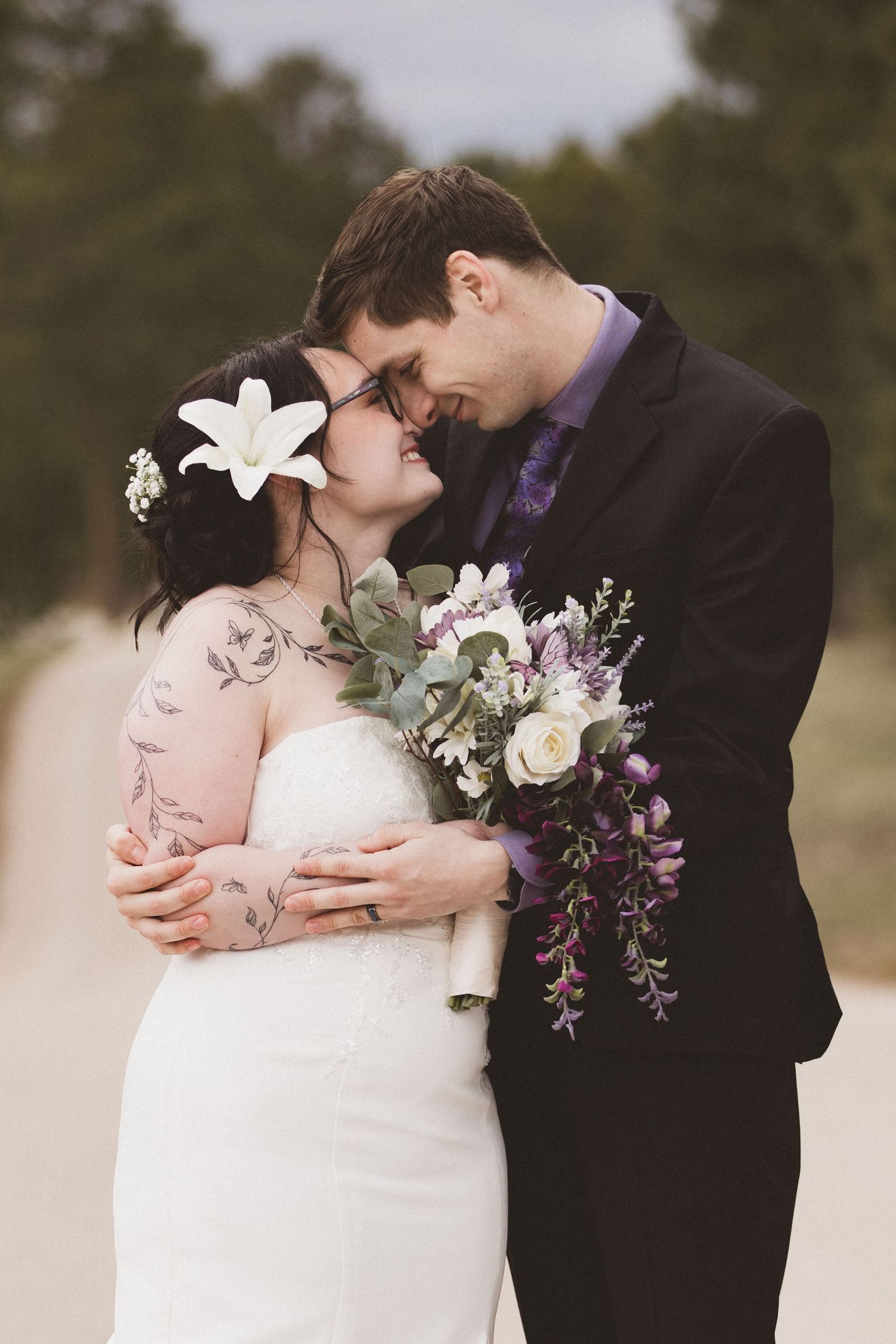 Bride and groom sharing romantic moment, she holds purple floral bouquet, white lily in hair, outdoor wedding portrait.