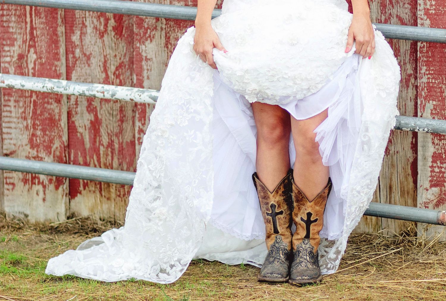 Bride in white lace wedding dress lifting skirt to reveal decorative cowboy boots, standing near rustic red barn fence.