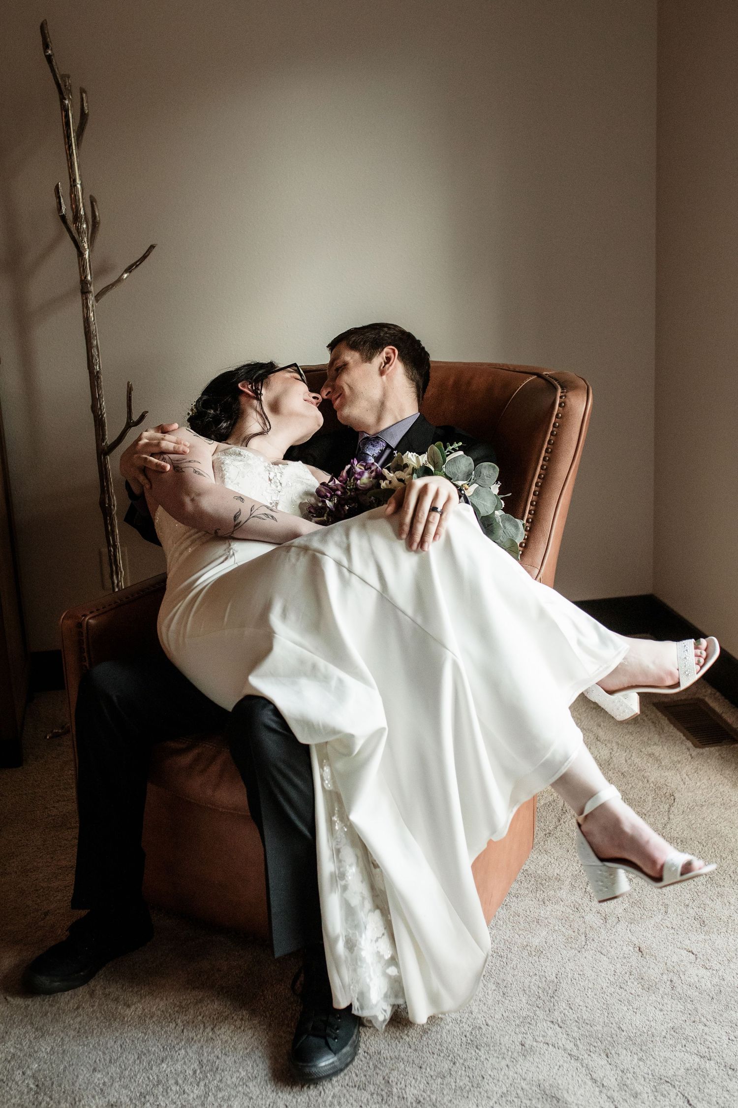 A groom holds his bride in a leather armchair as they share a romantic kiss in their wedding attire.