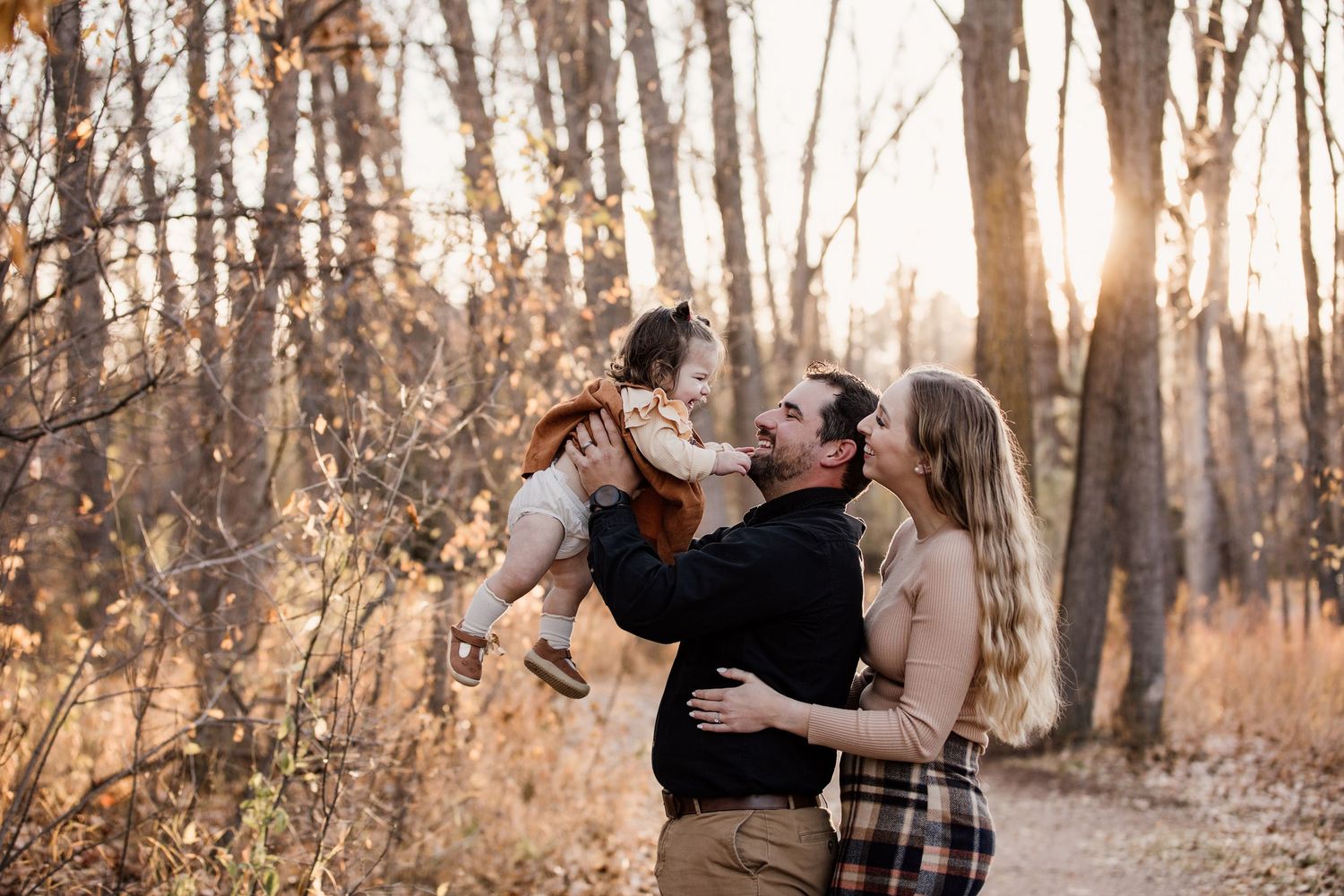 Family enjoying a golden sunset in autumn woods, parents lifting laughing toddler among fall foliage.