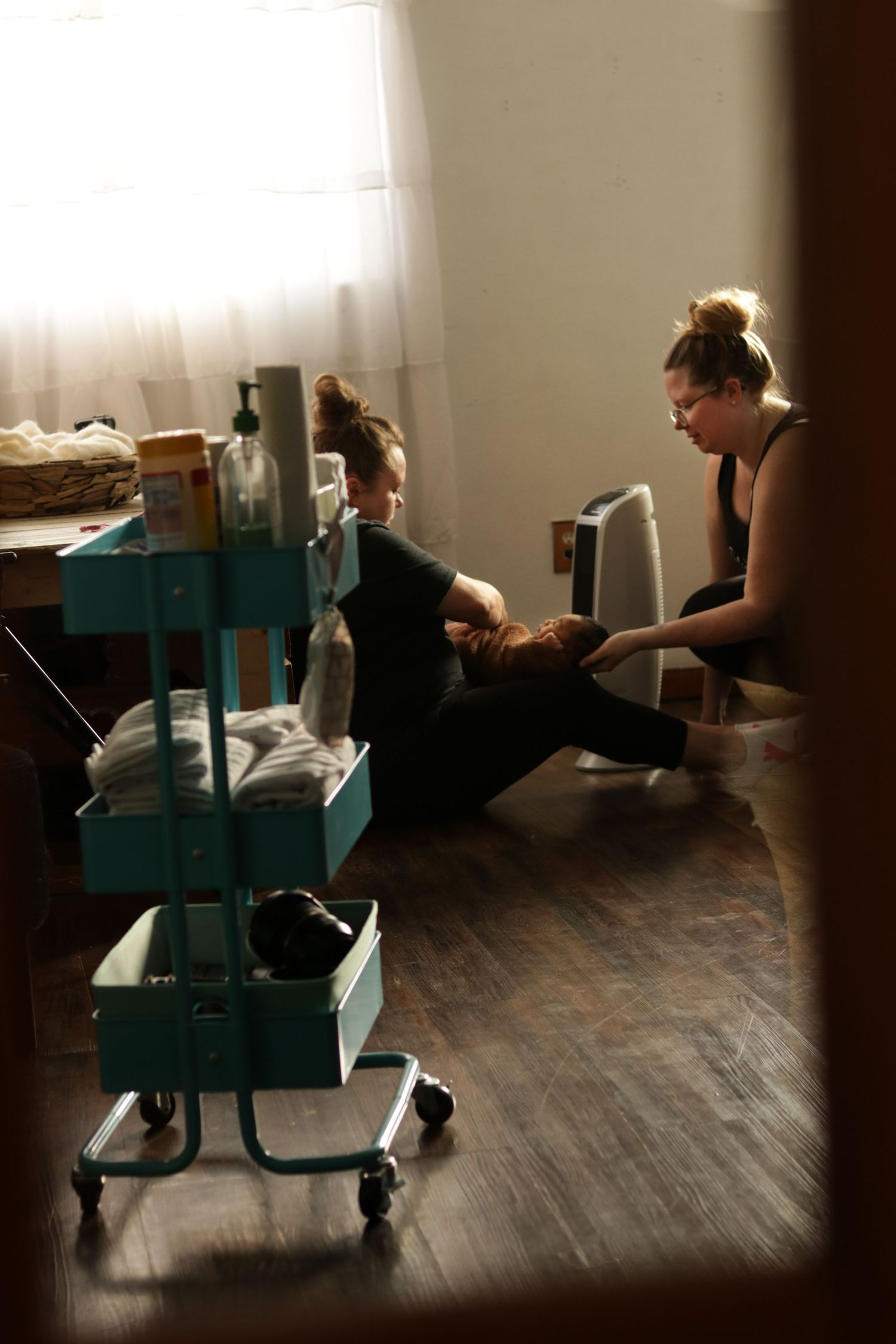 People sitting on a hardwood floor near a turquoise rolling cart in a dimly lit room with sunlight filtering through curtains.