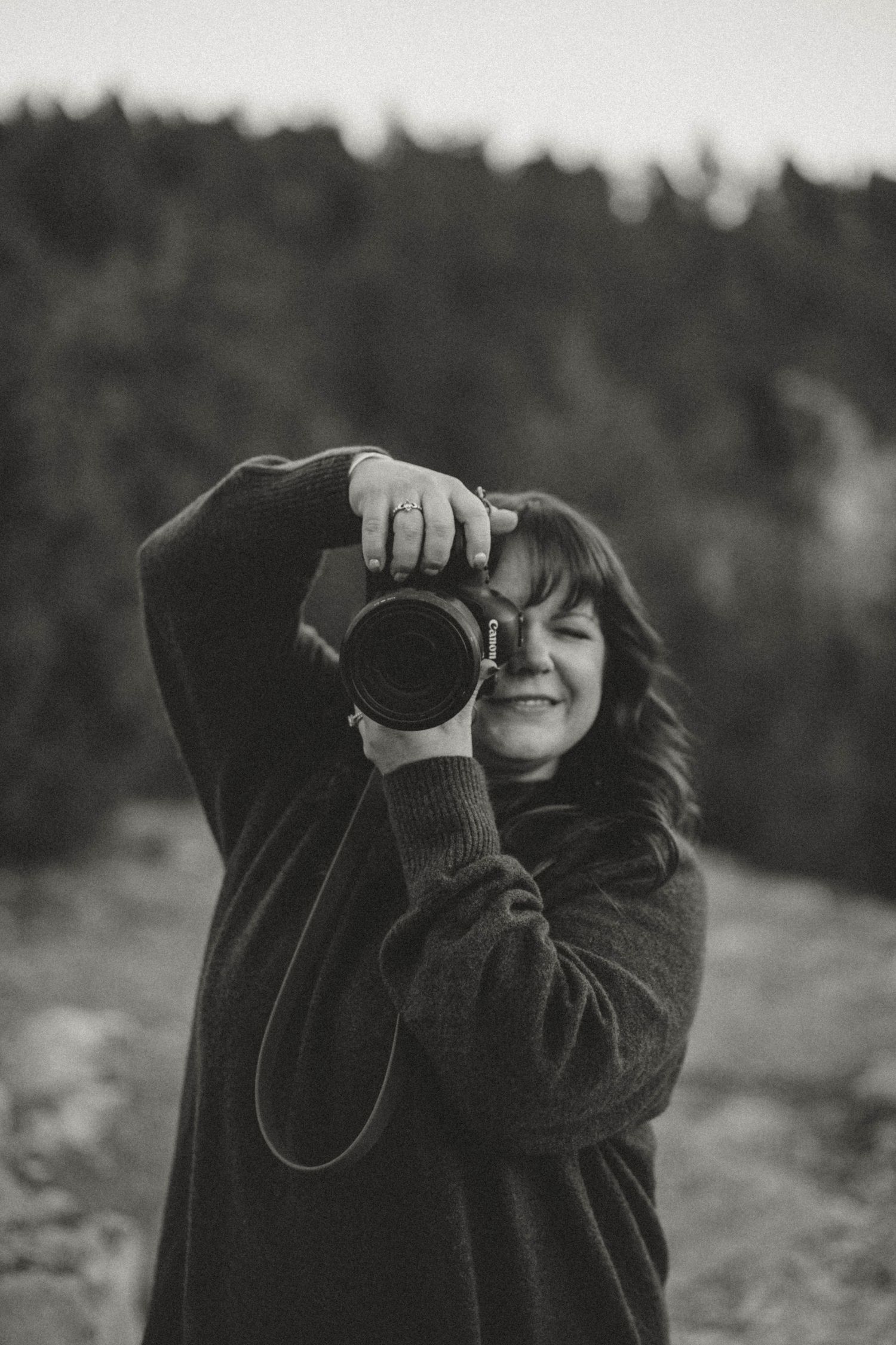 Black and white photo of a smiling woman holding a camera up to shoot in an outdoor forest setting.