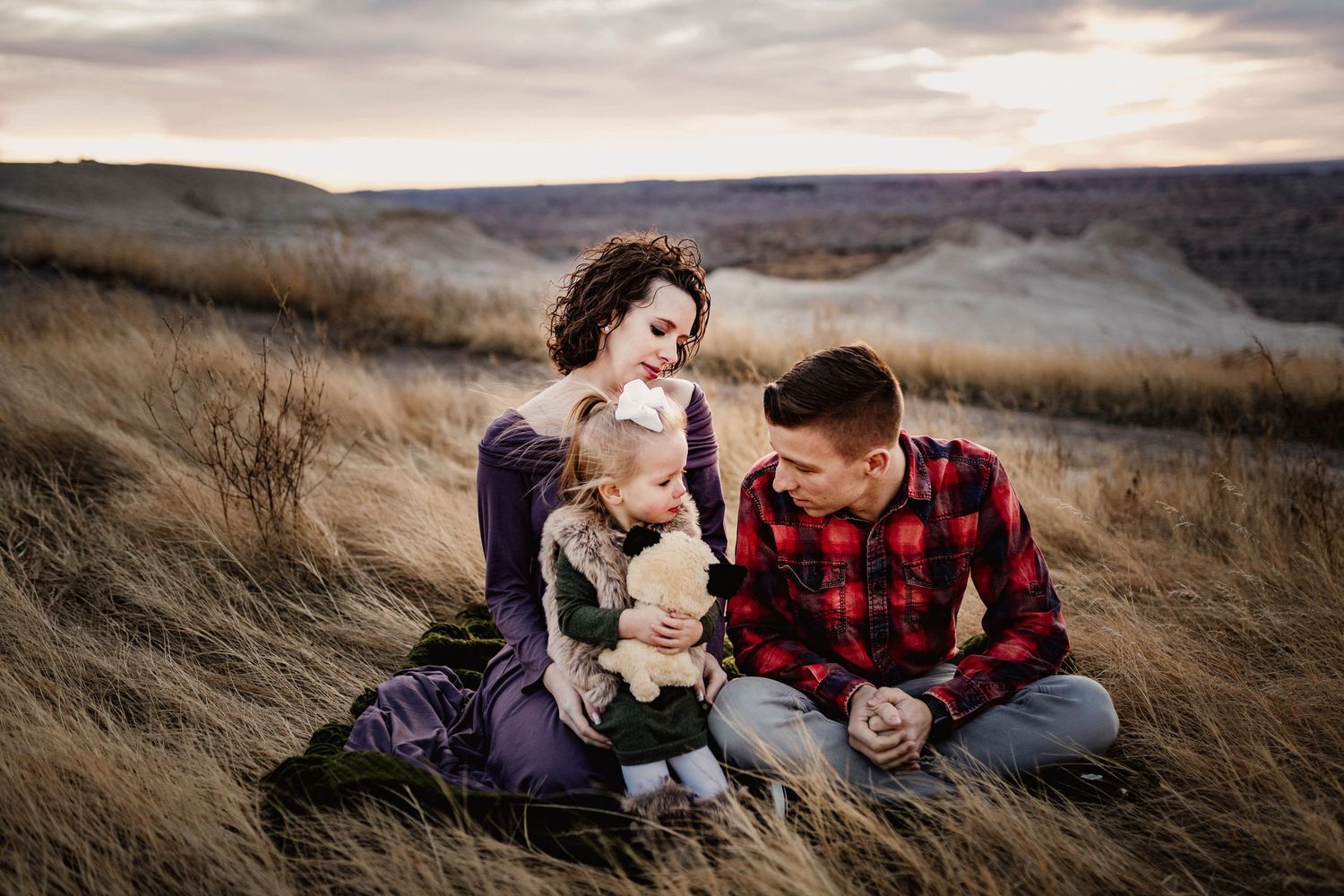 Family sitting in golden prairie grass with baby, woman in purple dress and man in red plaid shirt.