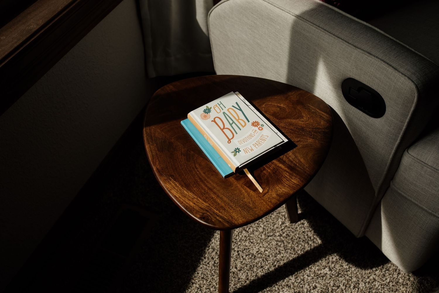 Baby book rests on a round wooden side table beside a gray chair in warm sunlight.