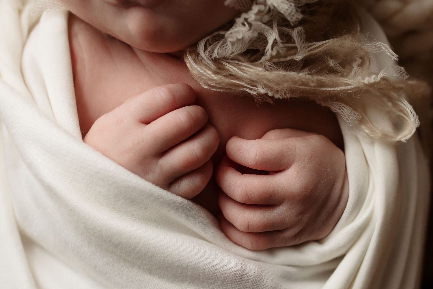 Close-up of newborn baby tiny hands and fingers wrapped in soft white swaddle cloth.