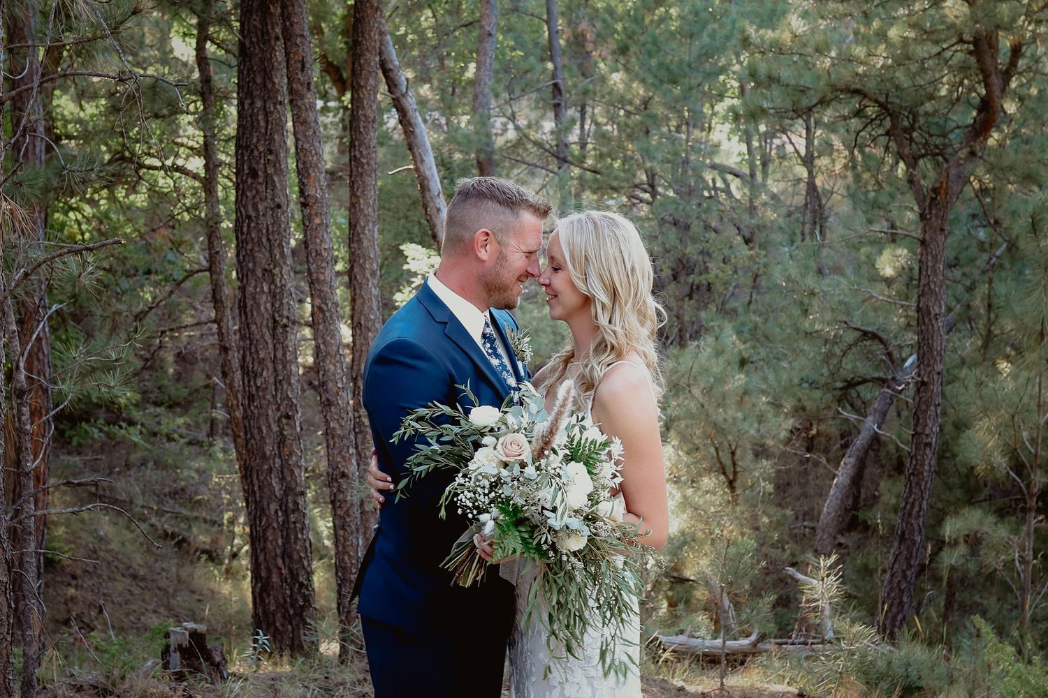 Bride and groom share a romantic forehead touch in a pine forest, bride holding a lush boho bouquet.