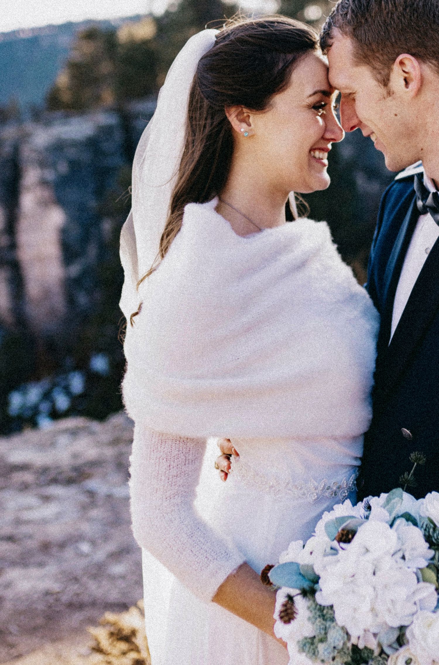 Bride in white fur wrap smiles at groom in navy suit, holding white bouquet, with rocky cliffs in background.