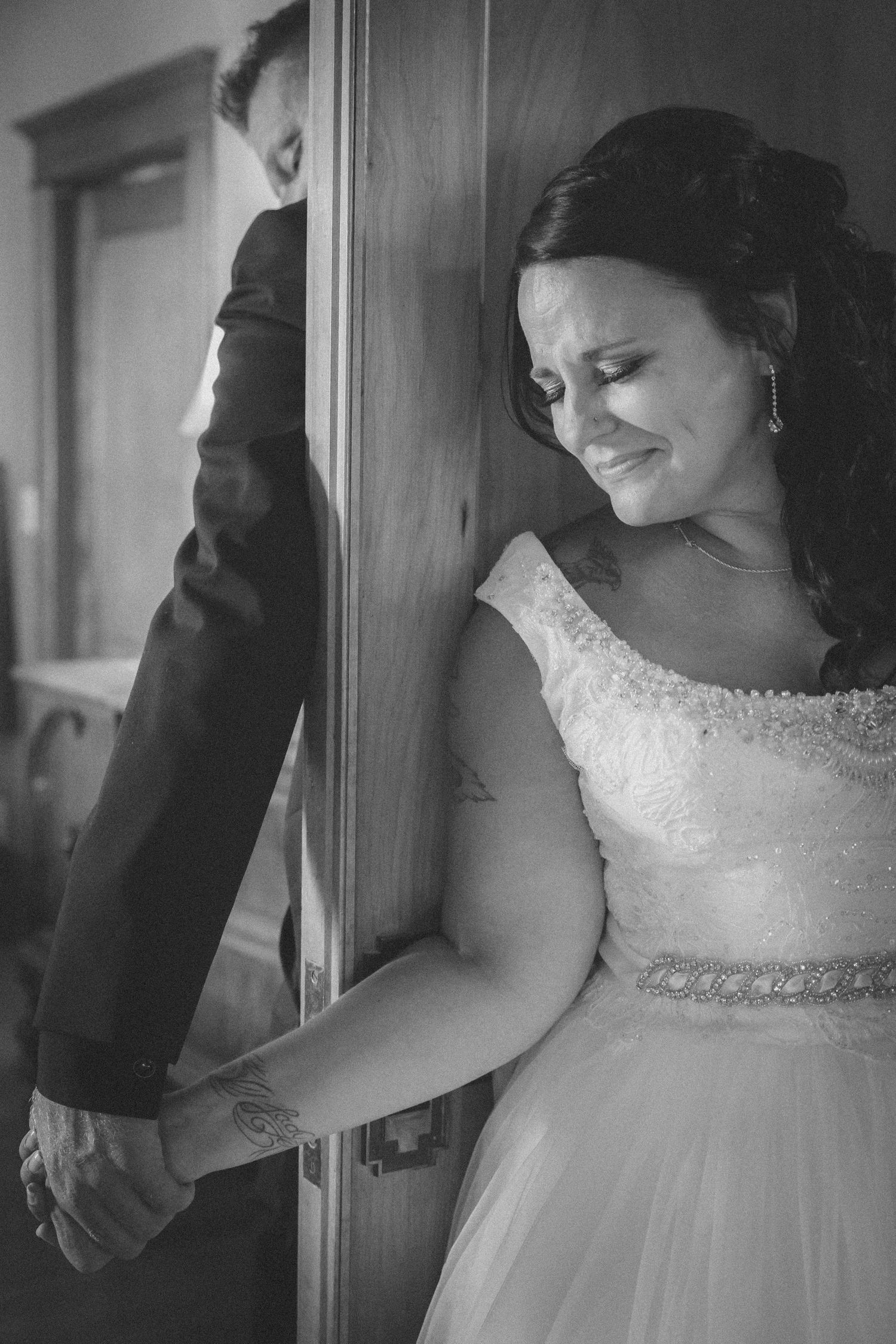 Black and white photo of emotional bride holding groom's hand through a door on their wedding day.